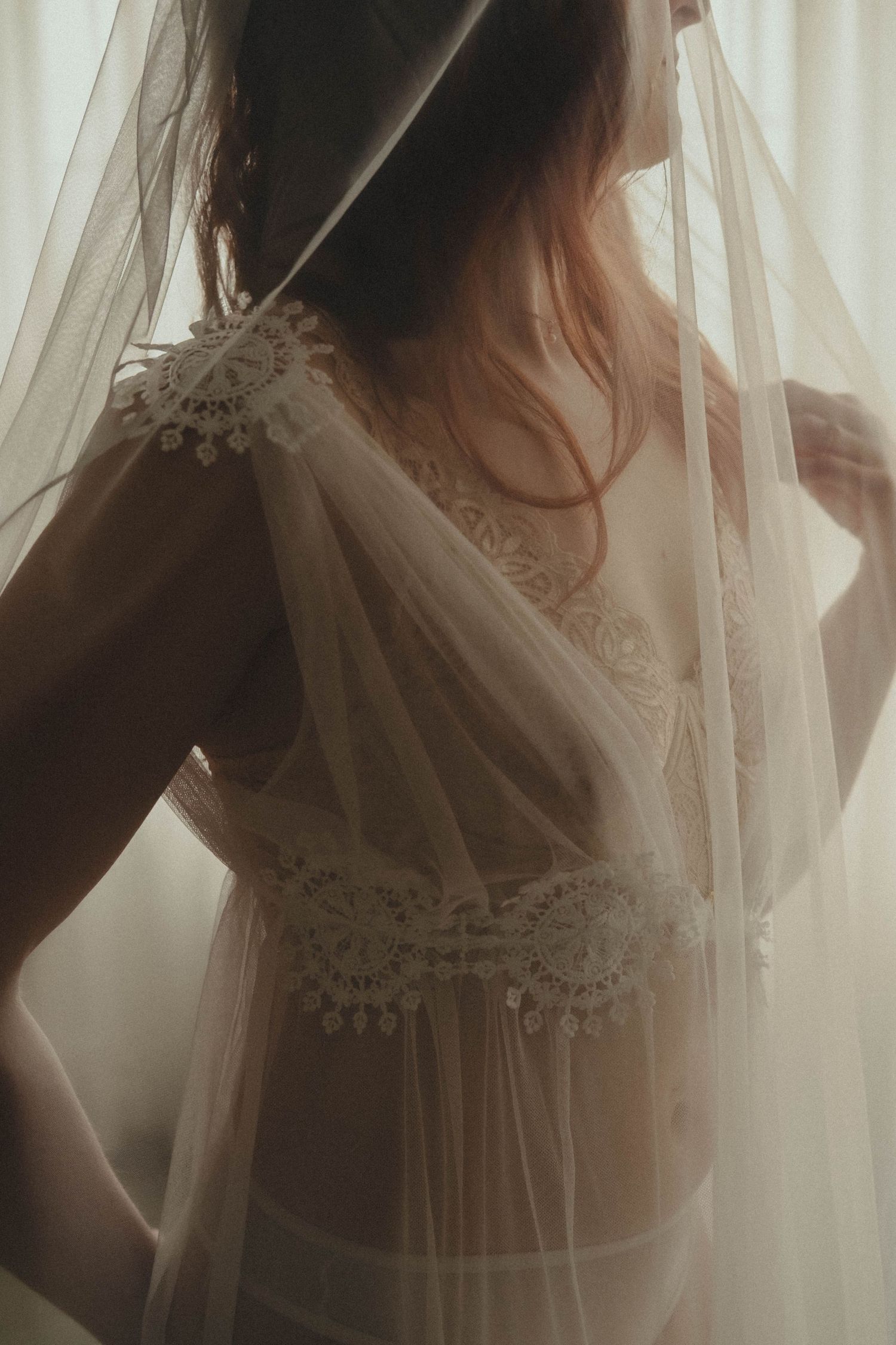 Bride in sheer white lace lingerie and veil, soft backlit boudoir portrait.