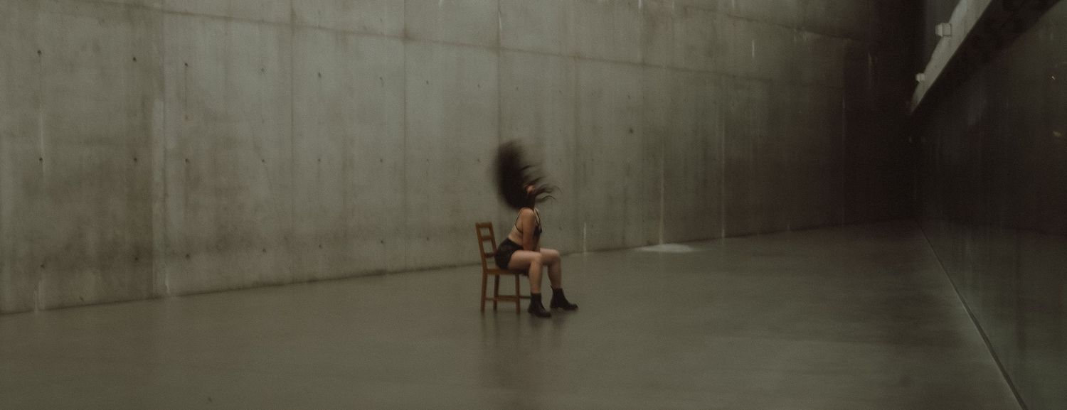 Person sitting on a wooden chair in a minimalist concrete room, head blurred in motion.