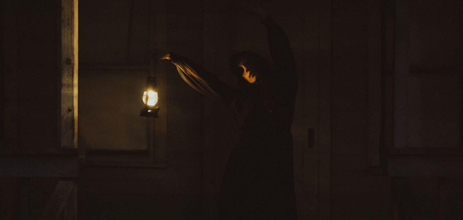 A mysterious silhouette holds a glowing lantern in a dark, dimly lit room with dramatic shadows.