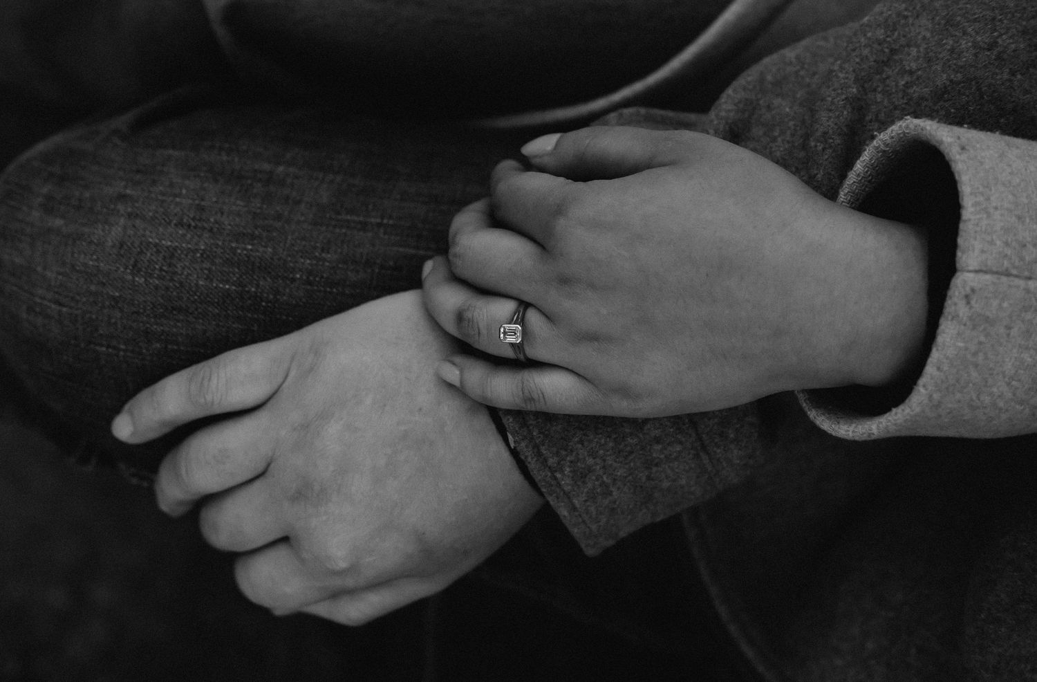 Black and white photo of two hands clasped together, one wearing a square diamond engagement ring.