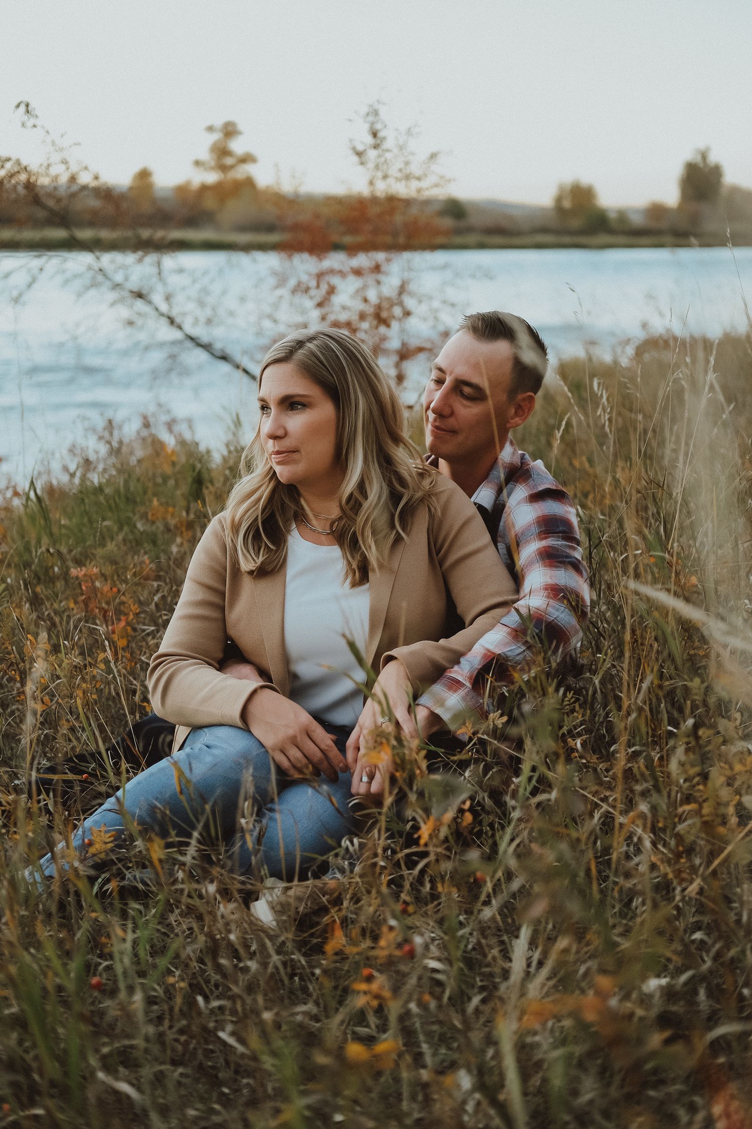 Couple sitting together in autumn tall grass by a lake during golden hour sunset.
