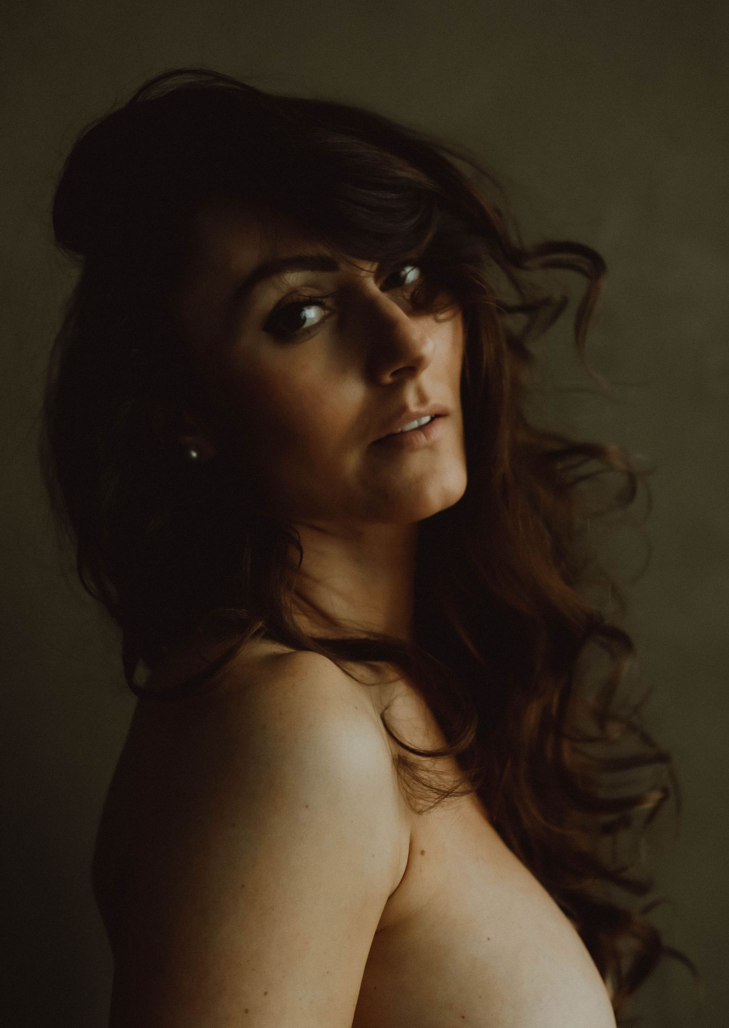 A woman with flowing auburn hair looks over her bare shoulder in moody, dramatic studio portrait photography.