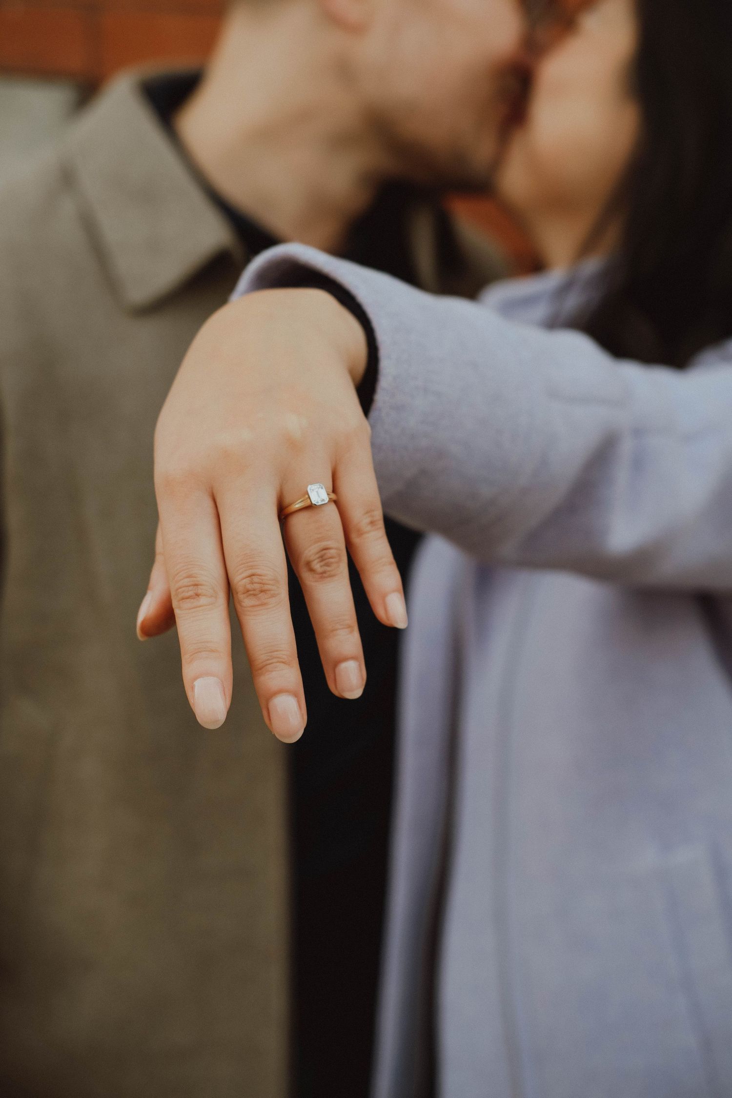 Couple sharing a kiss while woman displays her engagement ring with an emerald-cut diamond solitaire in the foreground.