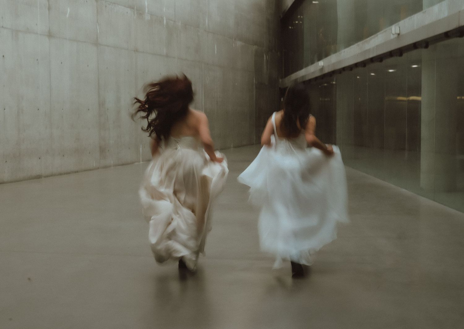 Two women in flowing white dresses running through a modern concrete building, motion blur capturing graceful movement.