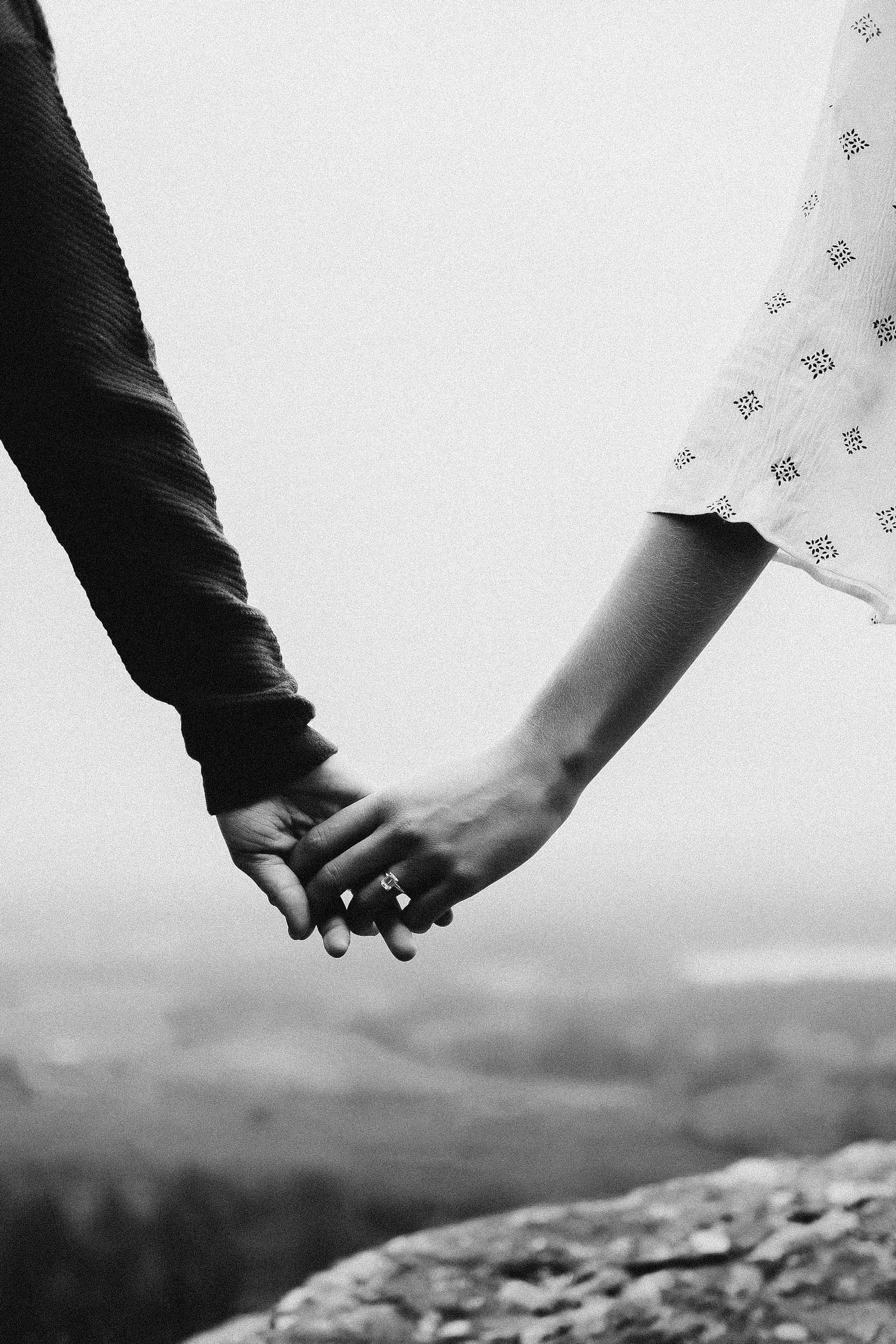Black and white photo of a couple holding hands on a rocky overlook, woman wearing a ring and patterned dress.