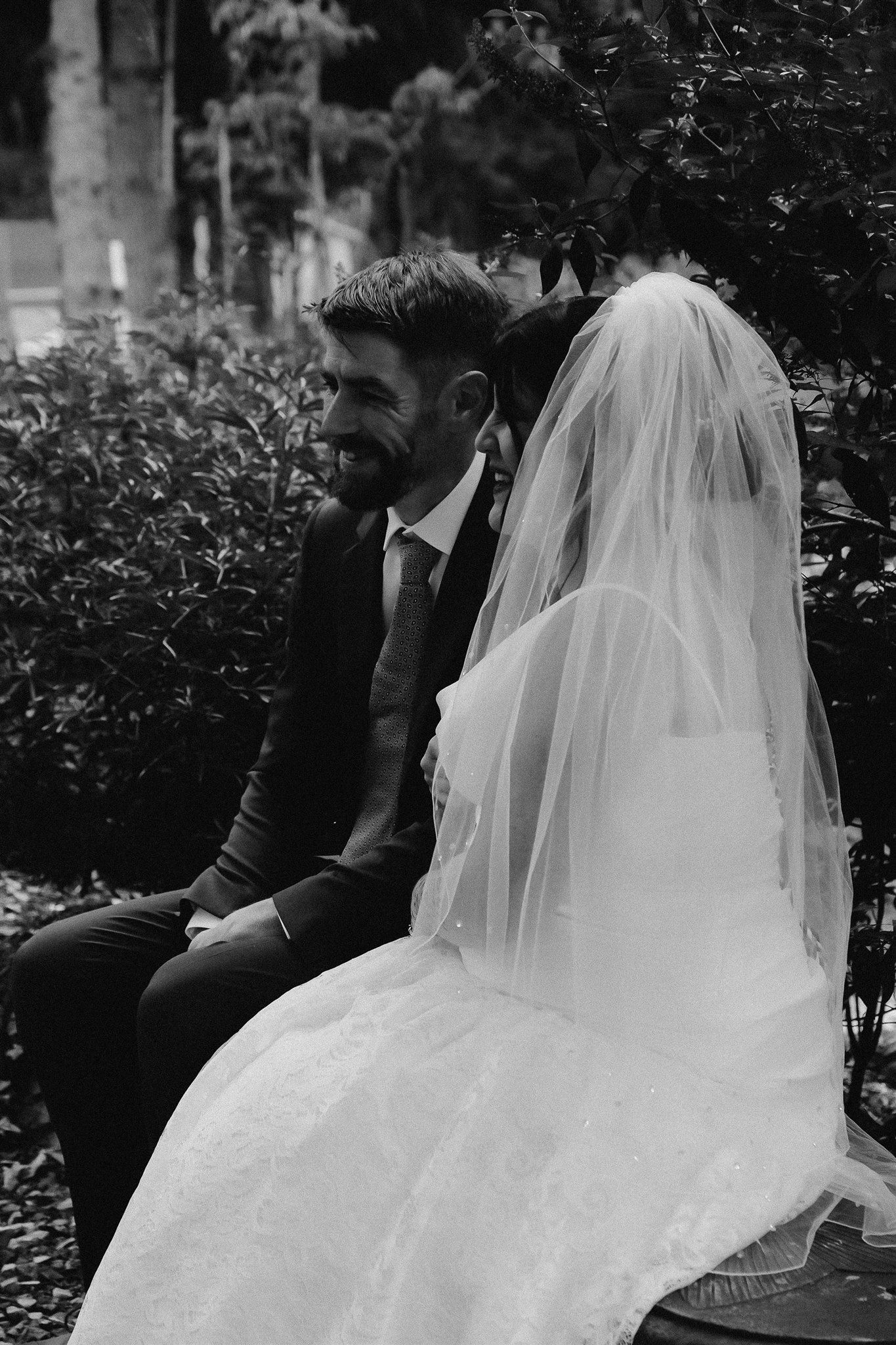 Smiling groom sitting beside veiled bride on bench in black and white garden portrait.