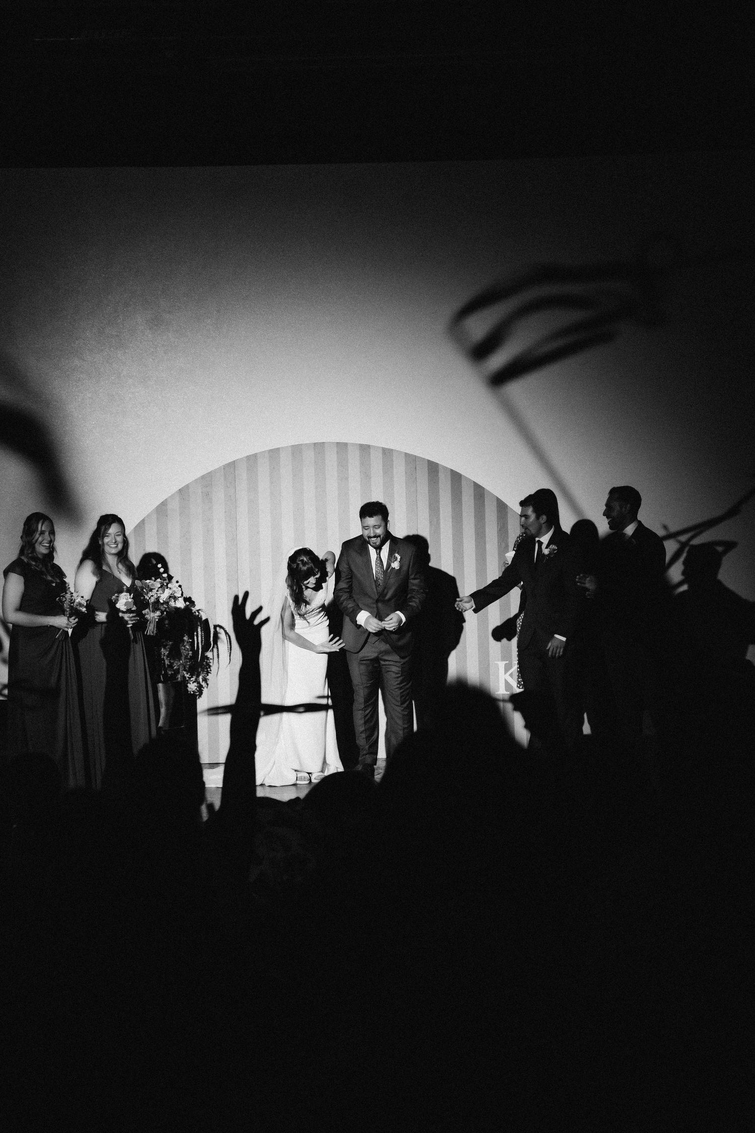 Black and white photo of bride and groom on stage surrounded by wedding party during indoor ceremony.