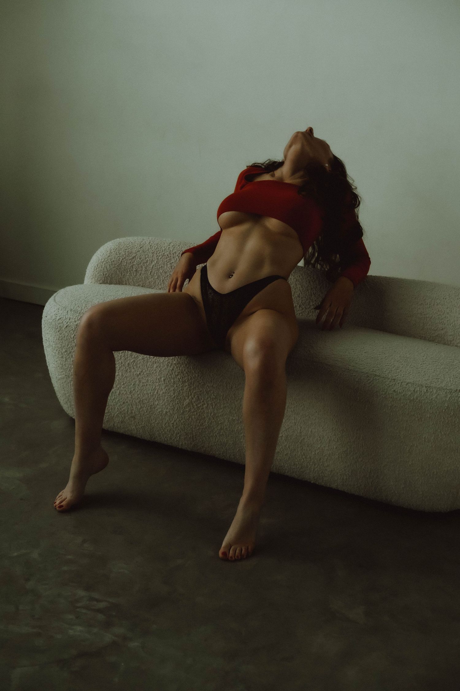 Woman in red top and dark underwear posing on a white boucle sofa in a minimalist studio setting.