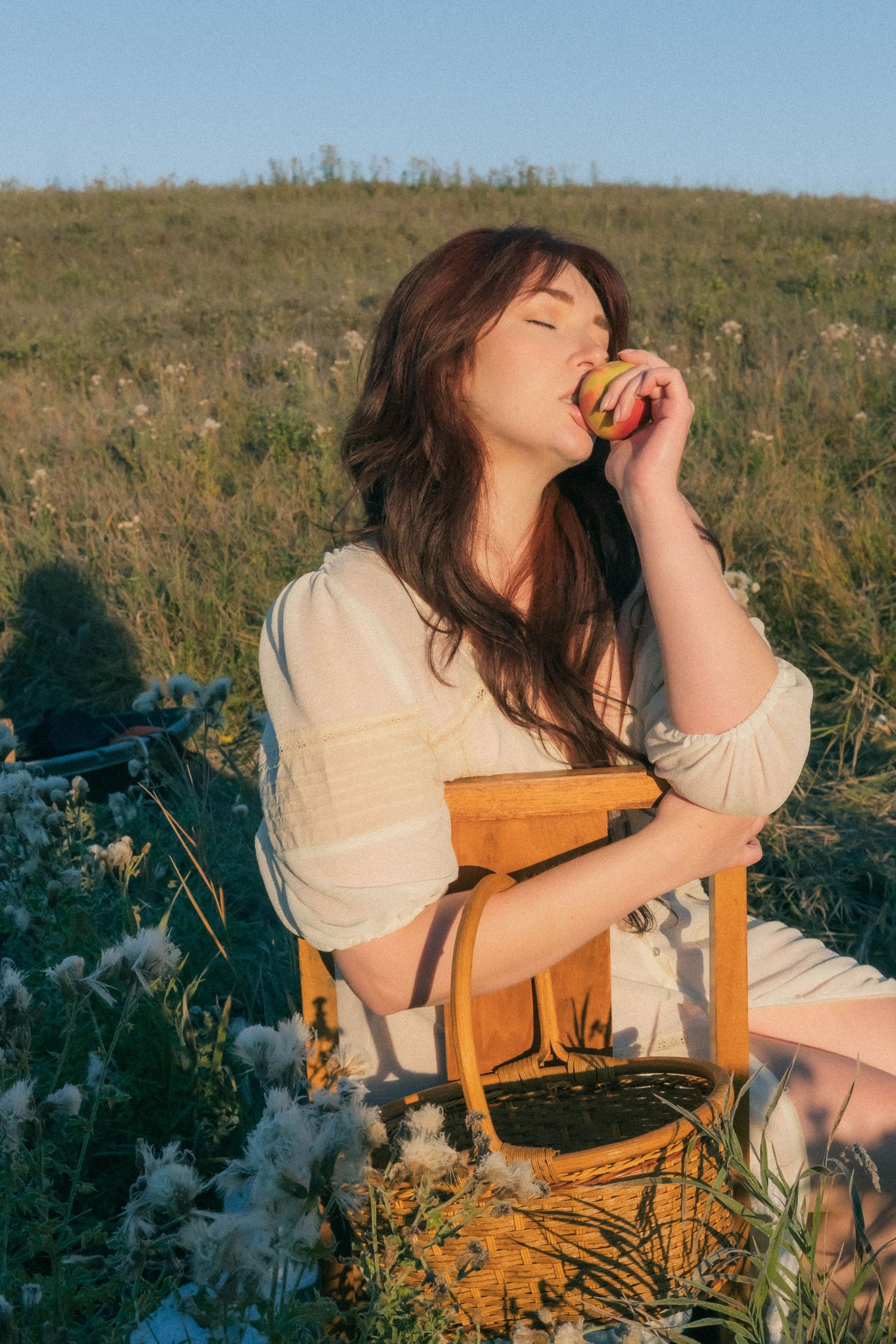 Young woman in white dress eating an apple while sitting on a wooden chair in a sunny green meadow.