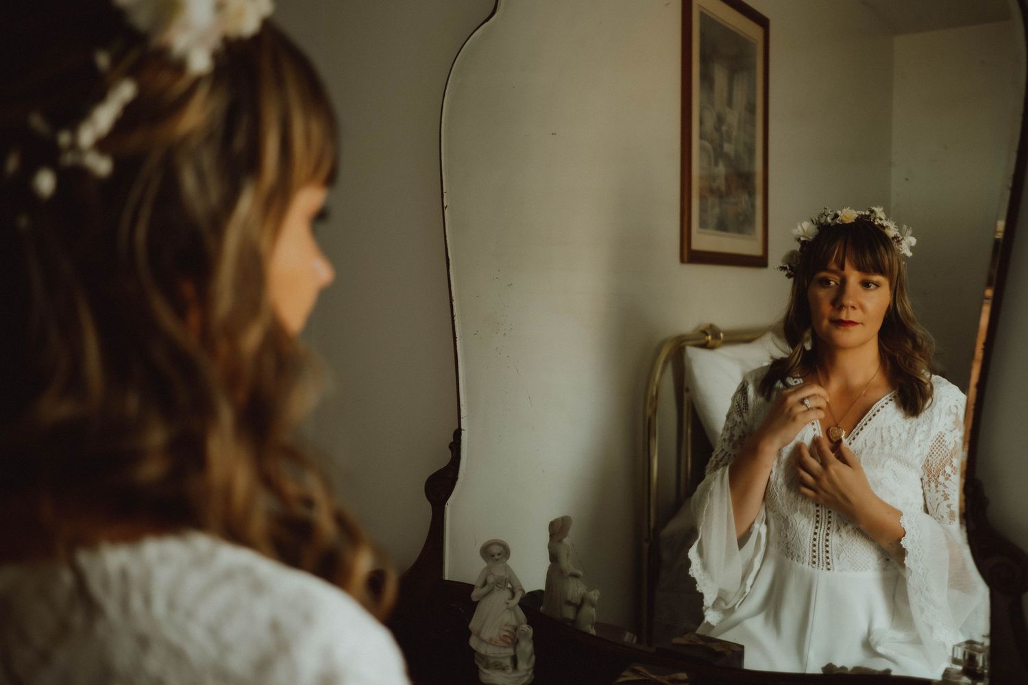Bride in white lace dress with floral crown prepares in front of vintage mirror.