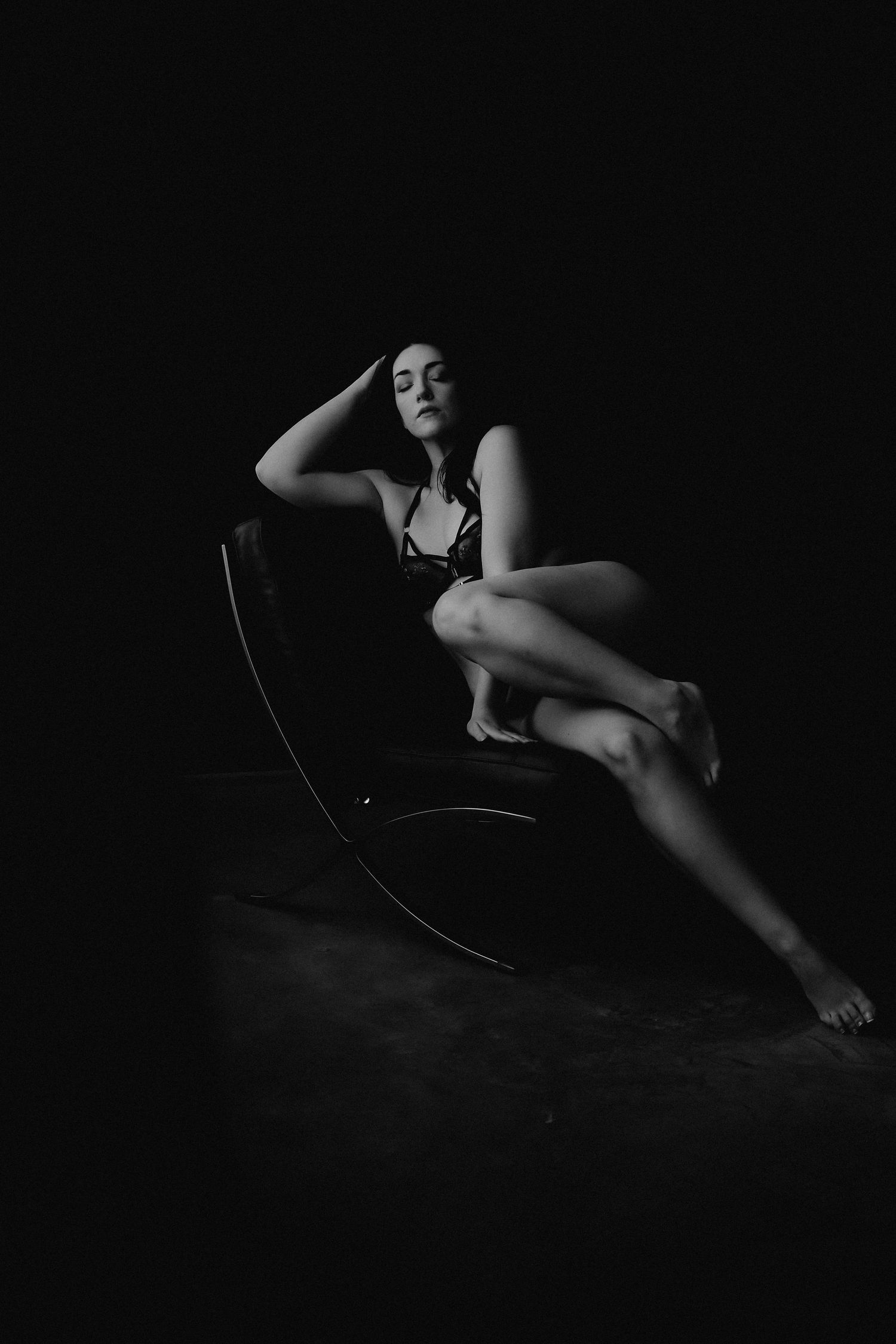 Black and white boudoir photo of a woman posing gracefully in a chair against a dark background.
