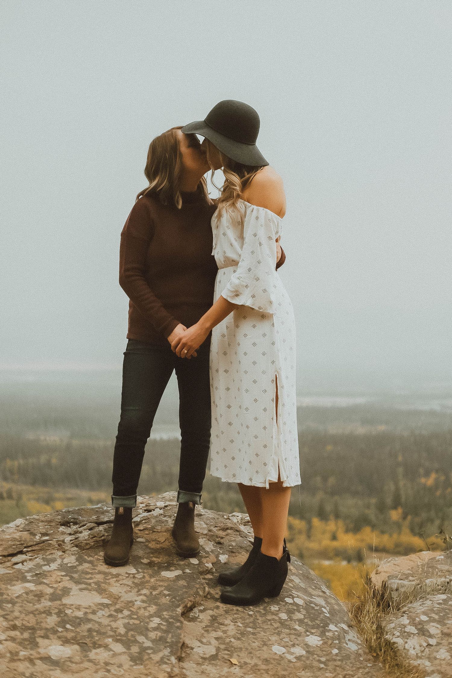 Two women sharing a kiss on a rocky overlook, one in a floral dress and wide-brim hat, holding hands with autumn views behind.