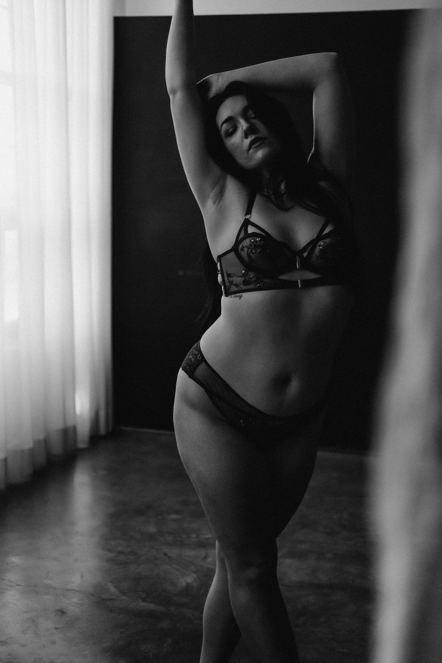 Black and white boudoir photo of a curvy woman posing in lace lingerie near a window with soft light.