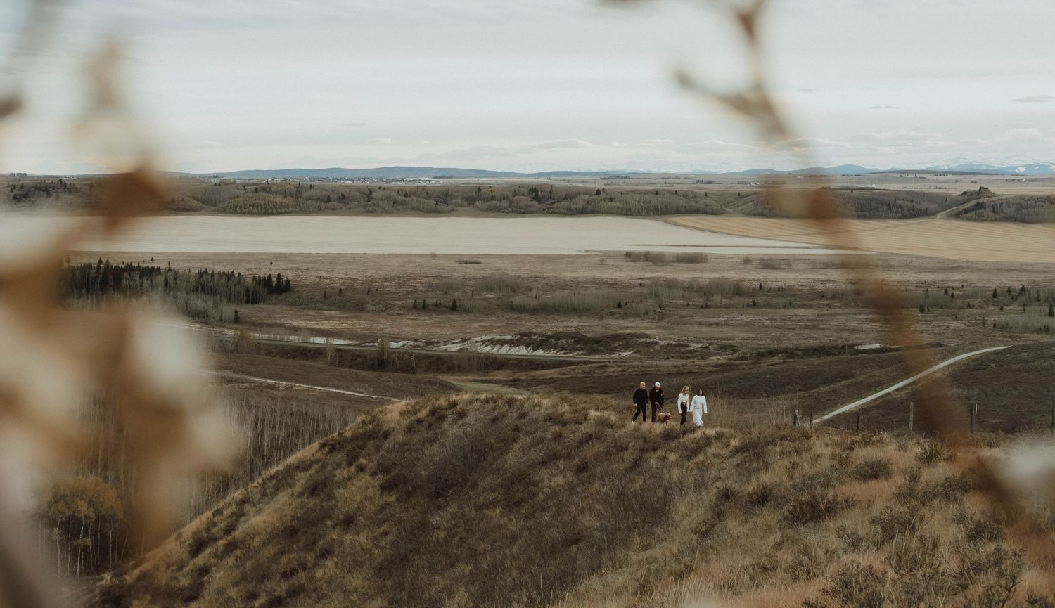 A small wedding party stands on a rolling prairie hillside overlooking a vast landscape with a lake in the distance.