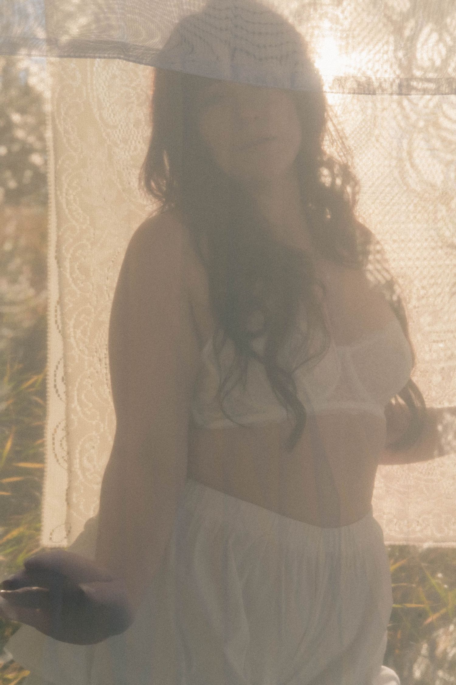 Dreamy soft-focus woman in white lingerie behind lace curtain outdoors.