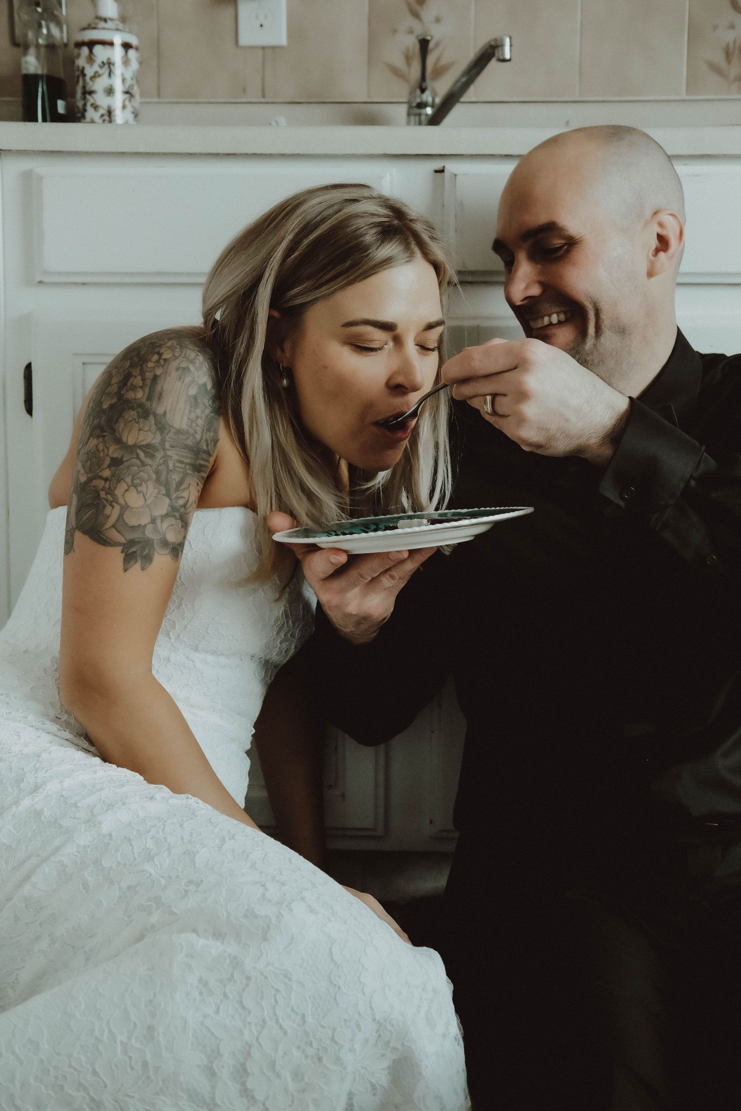 Bride in white dress with tattoos being fed cake by smiling bald groom in black suit at their wedding.