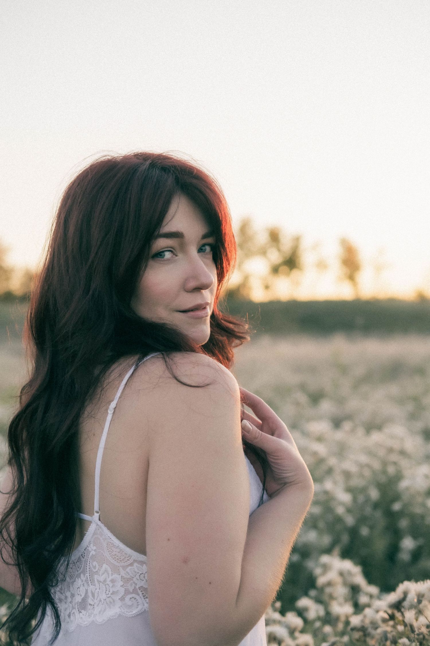 A young woman with long dark red hair poses in a sunlit wildflower field wearing a white lace dress at golden hour.