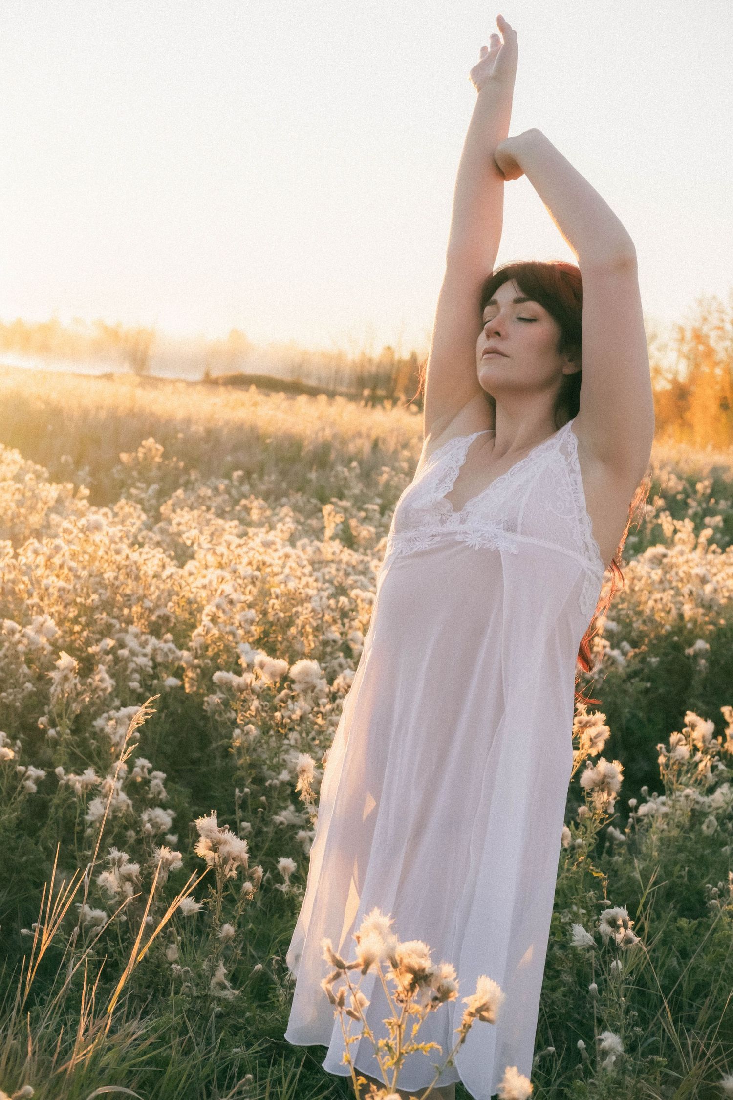Woman in white slip dress with arms raised in a sunlit wildflower field at dusk.