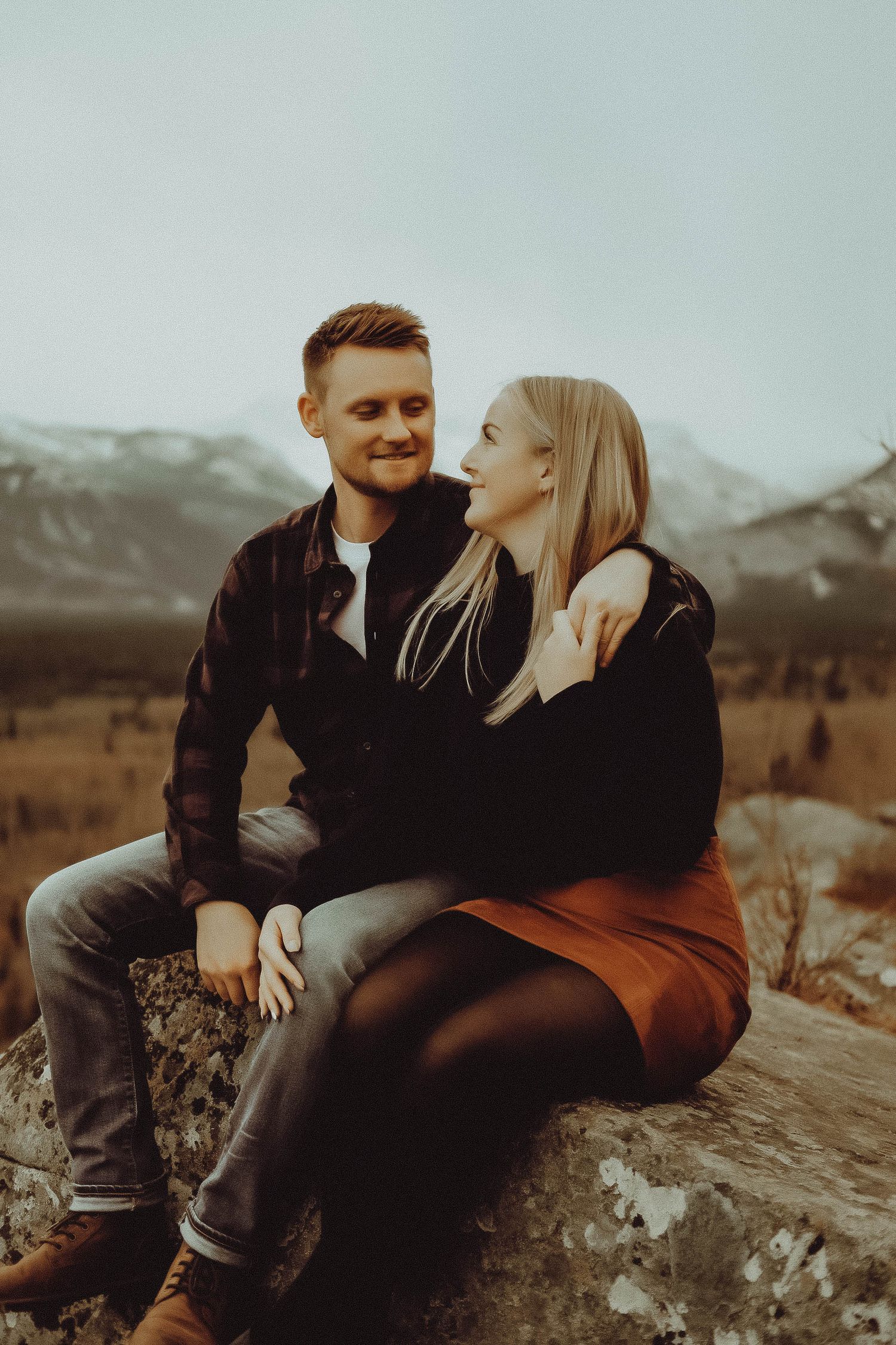 A smiling couple sits together on a rock with snow-capped mountains in the background.