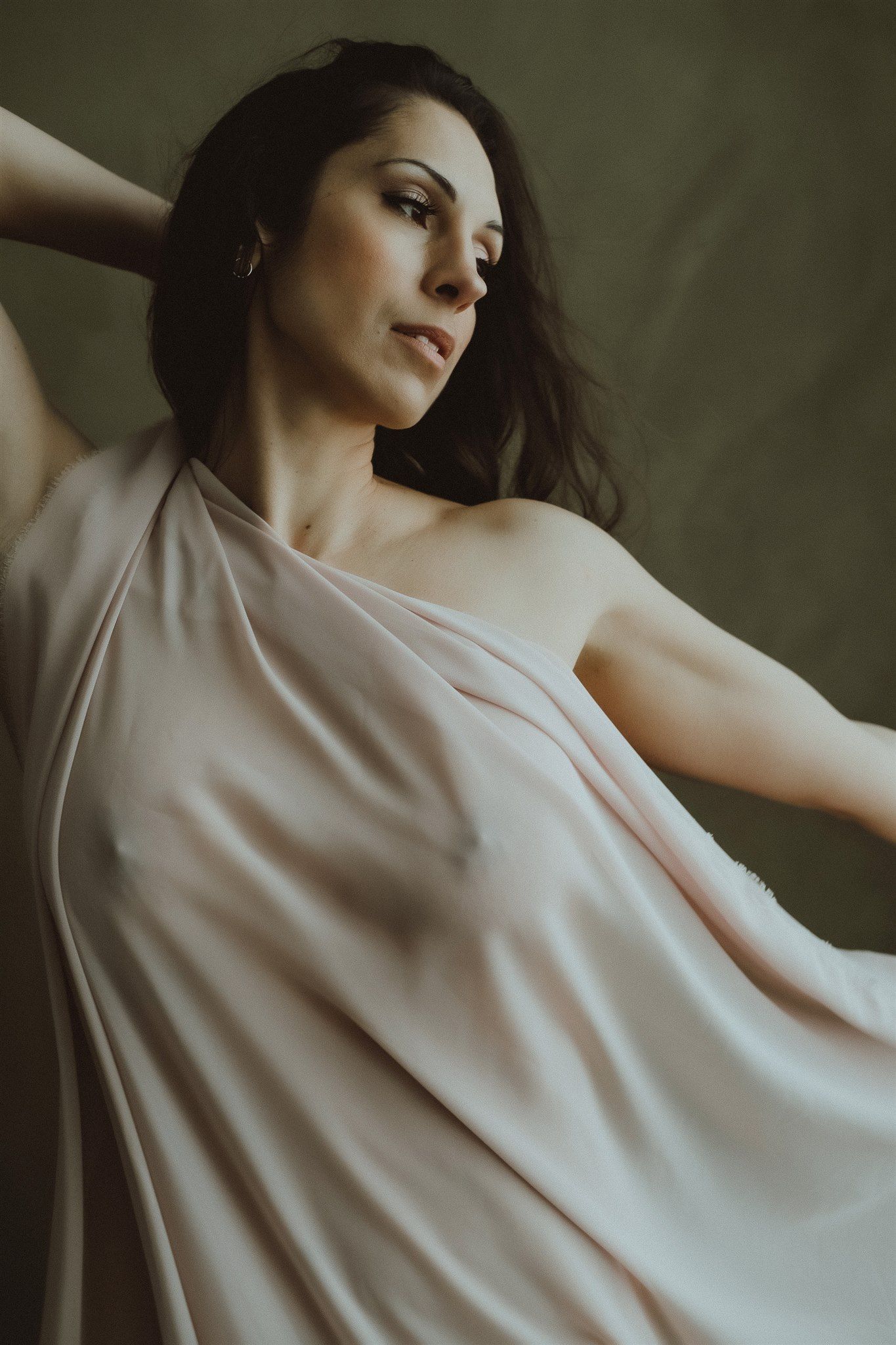 Elegant woman draped in flowing blush pink fabric posing against an olive green background.