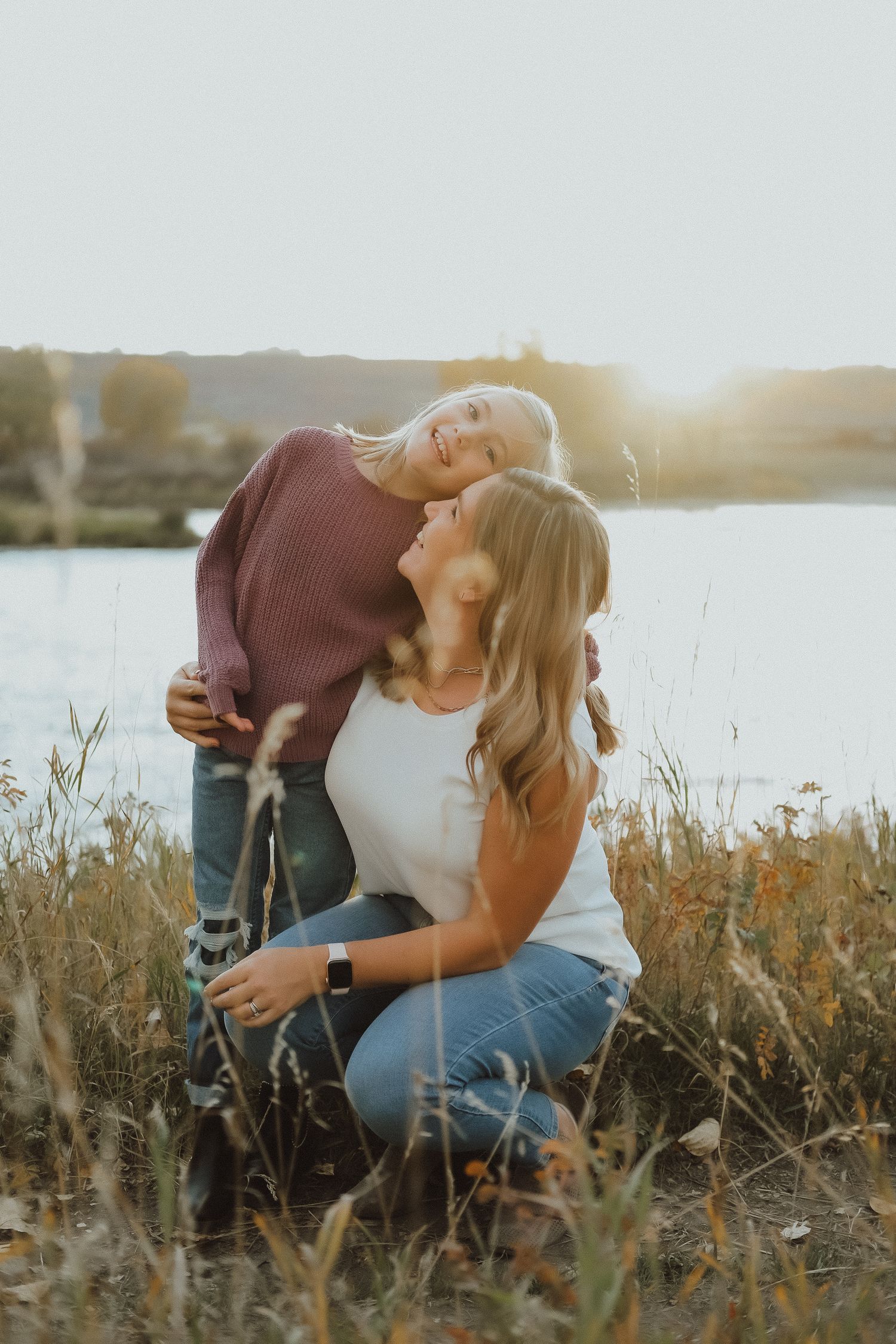 Mother and daughter sharing a tender moment in a sunlit field near a lake at golden hour.