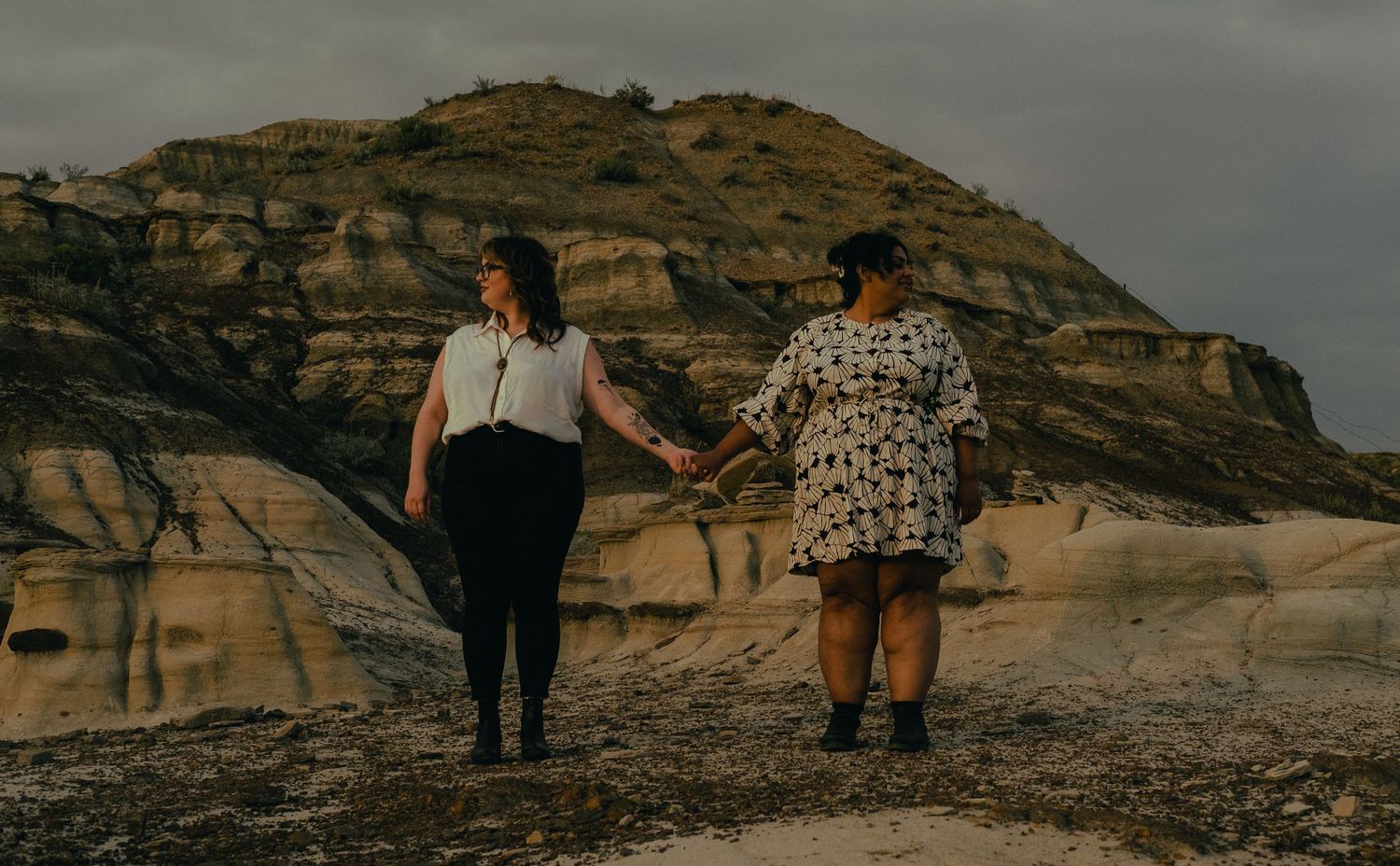 Two women holding hands while walking through a dramatic badlands landscape with rocky hoodoos.