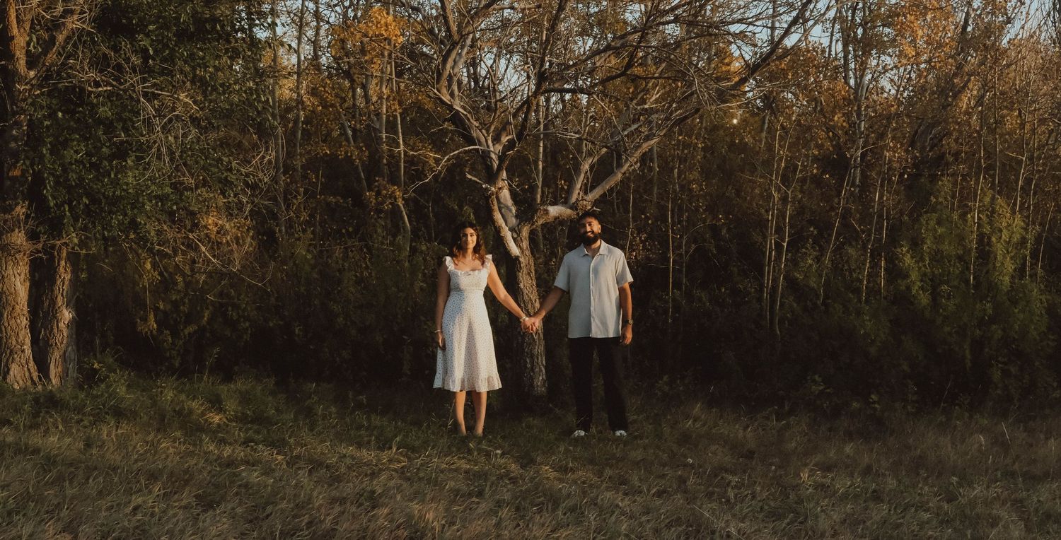 Couple holding hands in a sunlit forest clearing, woman in white dress, man in white shirt, golden hour lighting.