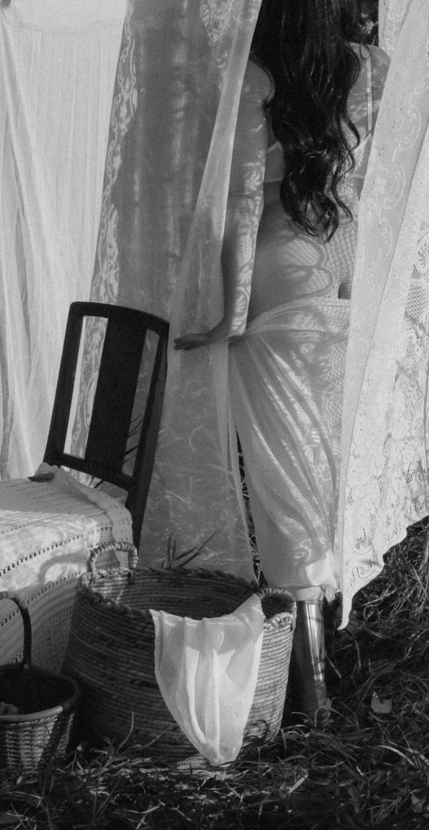 Black and white photo of a woman with curly hair standing among hanging white lace curtains with baskets and a chair nearby.