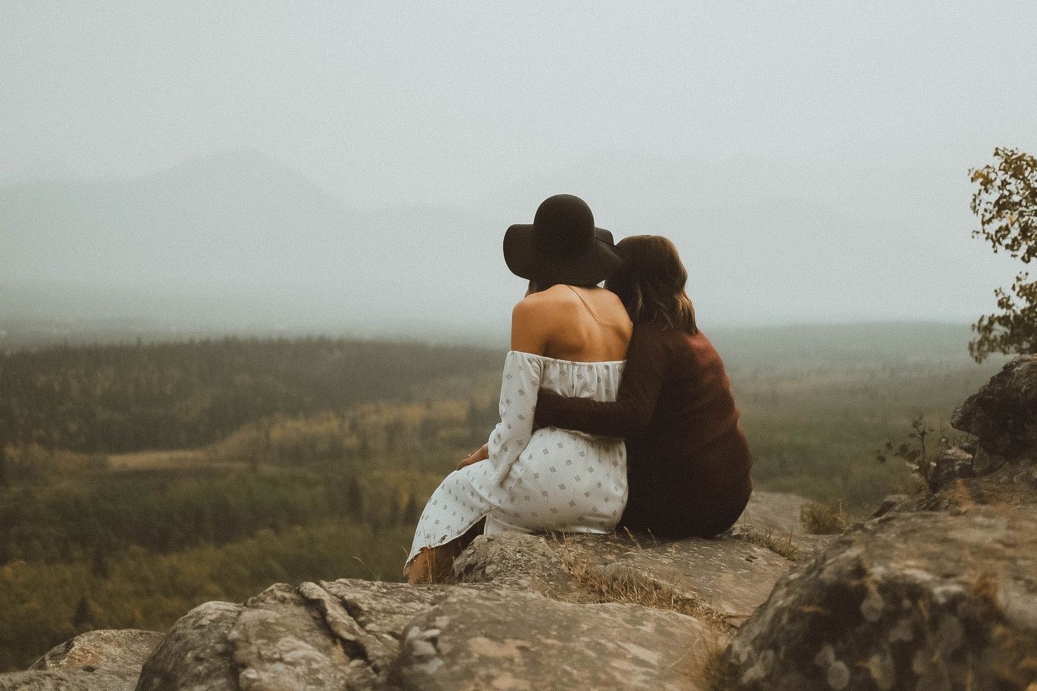 Two women sitting on a rocky cliff edge, embracing while overlooking a vast misty valley and mountain landscape.