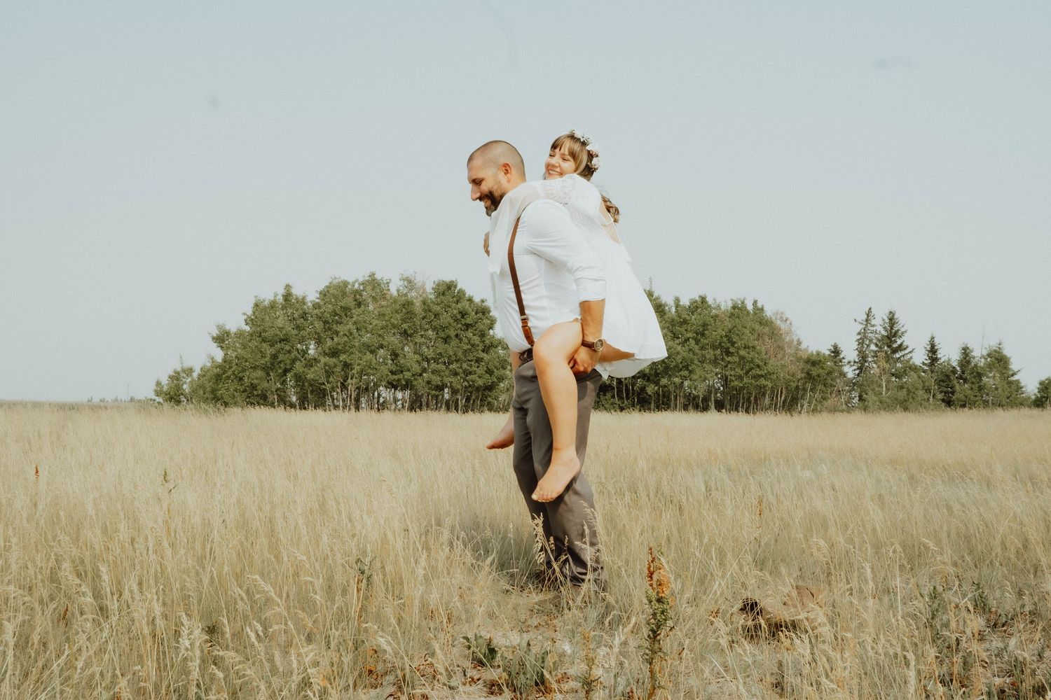 A man carries a woman on his back in a golden wheat field with trees in the background on a clear day.