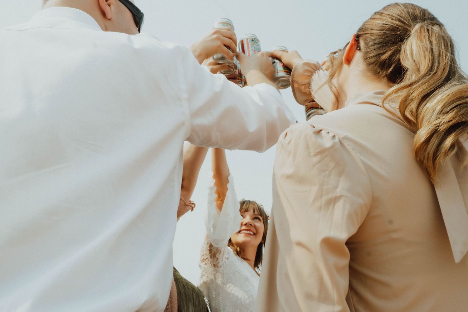 Group of friends in white shirts raising glasses in a celebratory toast outdoors under a bright sky.