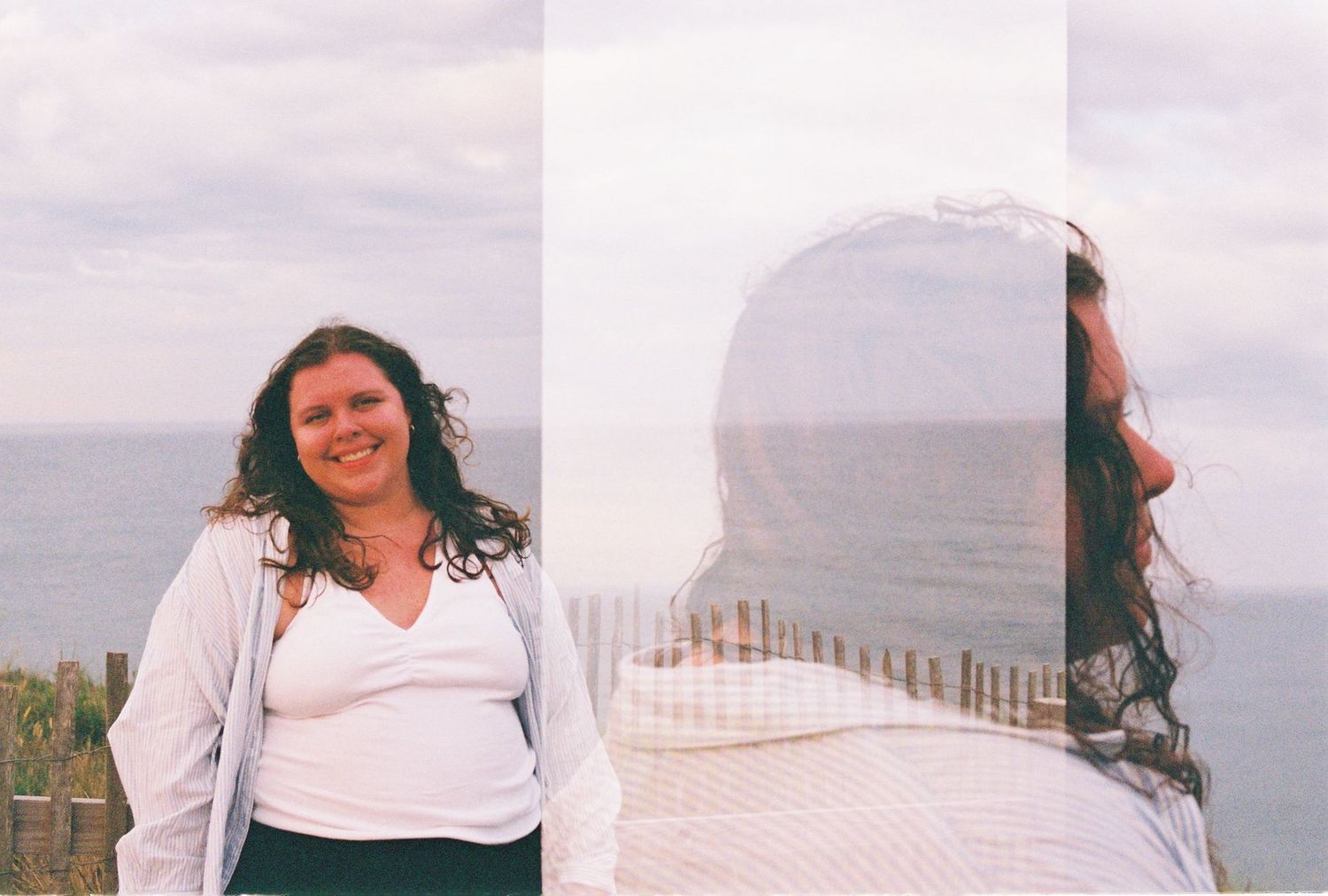 Double exposure photo of a smiling woman with curly hair standing by the ocean on a cloudy day.