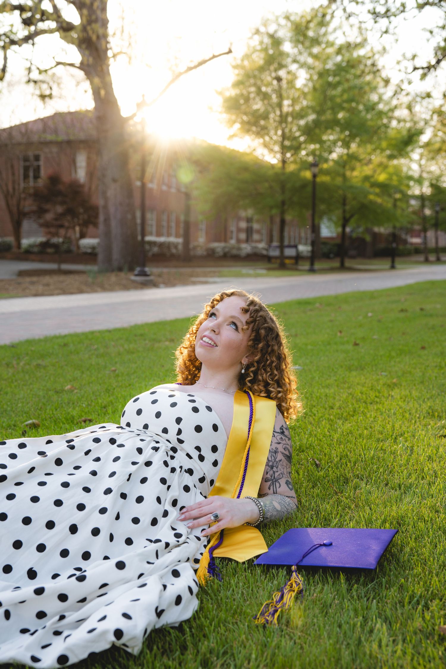 A curly-haired graduate in yellow sash and polka dot dress reclines on green grass at sunset with purple cap nearby.