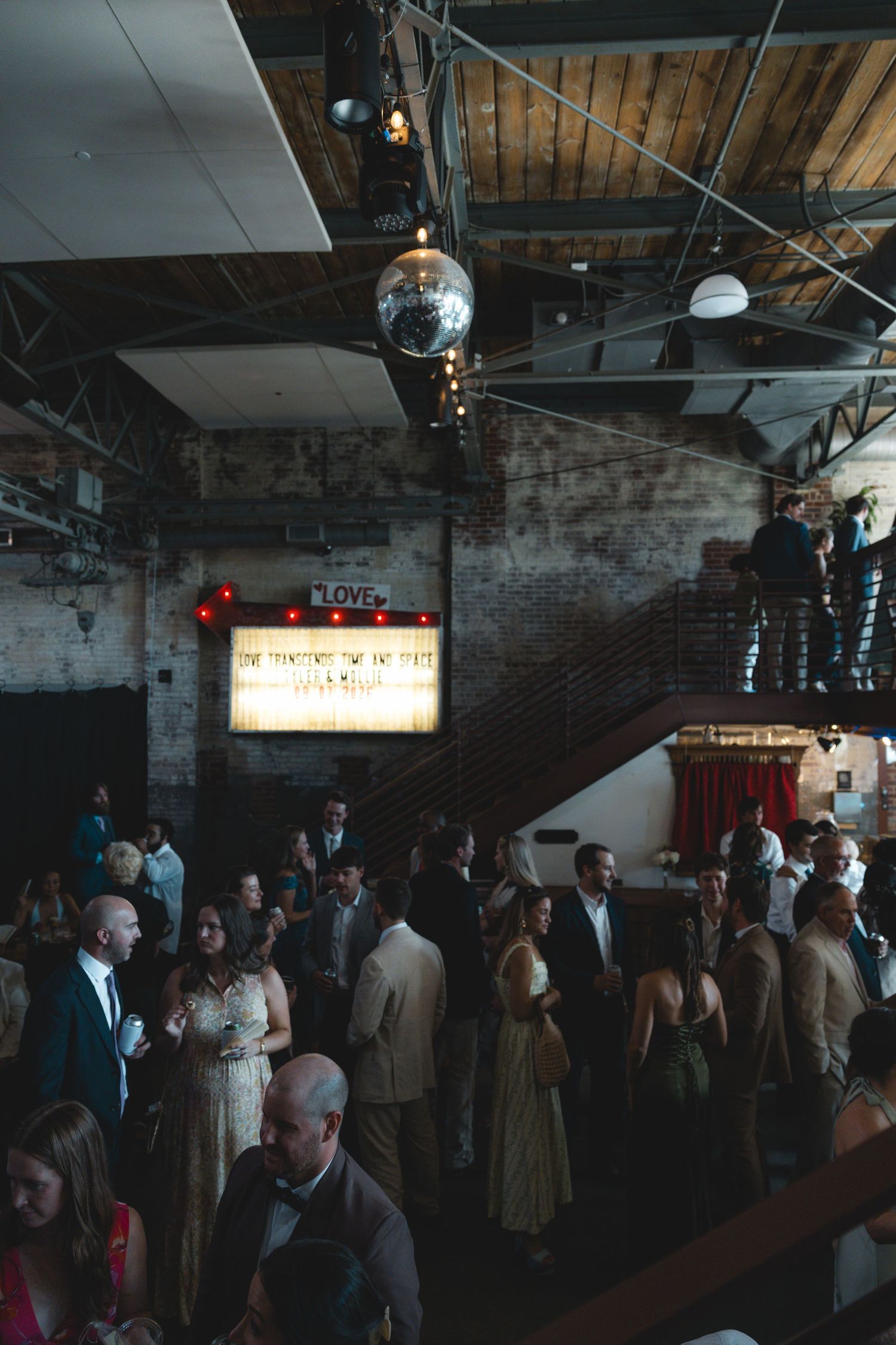 Guests mingle at an industrial wedding venue with disco ball, brick walls, and illuminated marquee sign.