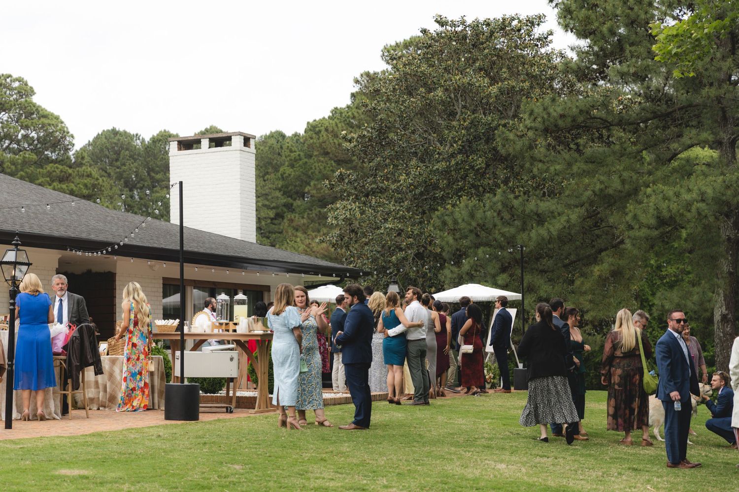 Guests mingle on green lawn outside rustic venue with white chimney and trees at an outdoor garden party.