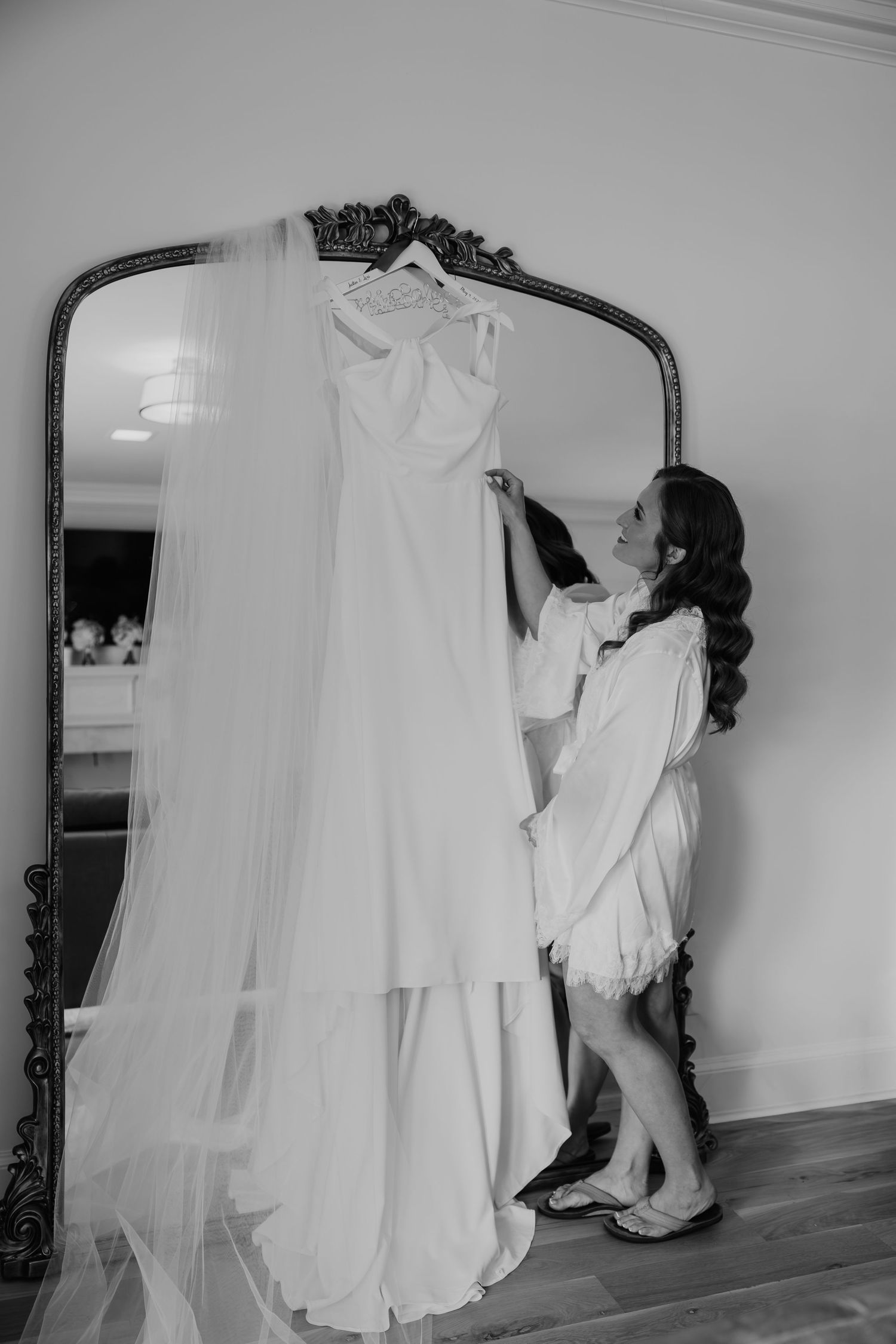 Bride in robe admiring her wedding dress and veil hanging on an ornate mirror in black and white photo.