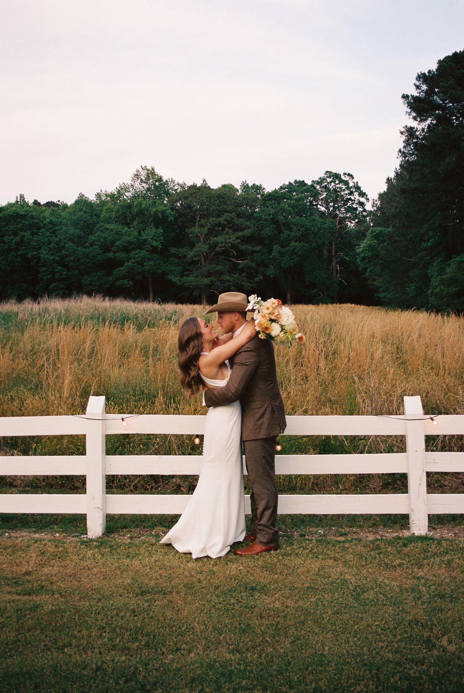Newlywed couple sharing a kiss by a white fence with golden fields and lush green trees in the background.