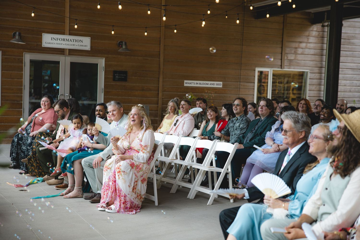 Guests seated in white chairs at an outdoor wedding ceremony venue with string lights and rustic barn backdrop.