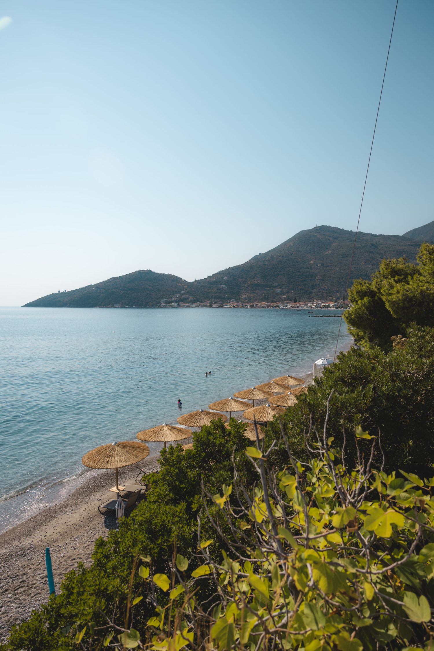 Serene Mediterranean beach with straw umbrellas, lush greenery, calm blue sea, and a hillside town in the background.