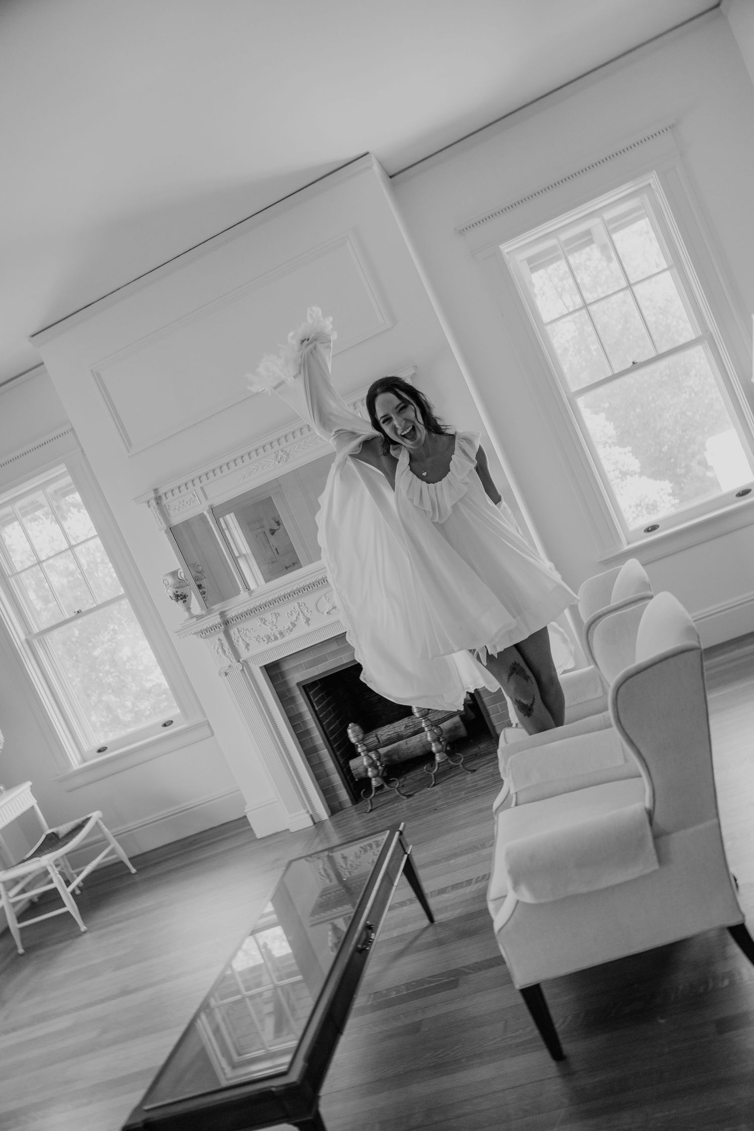Joyful bride in white robe dancing in elegant room with fireplace, captured in black and white photography.