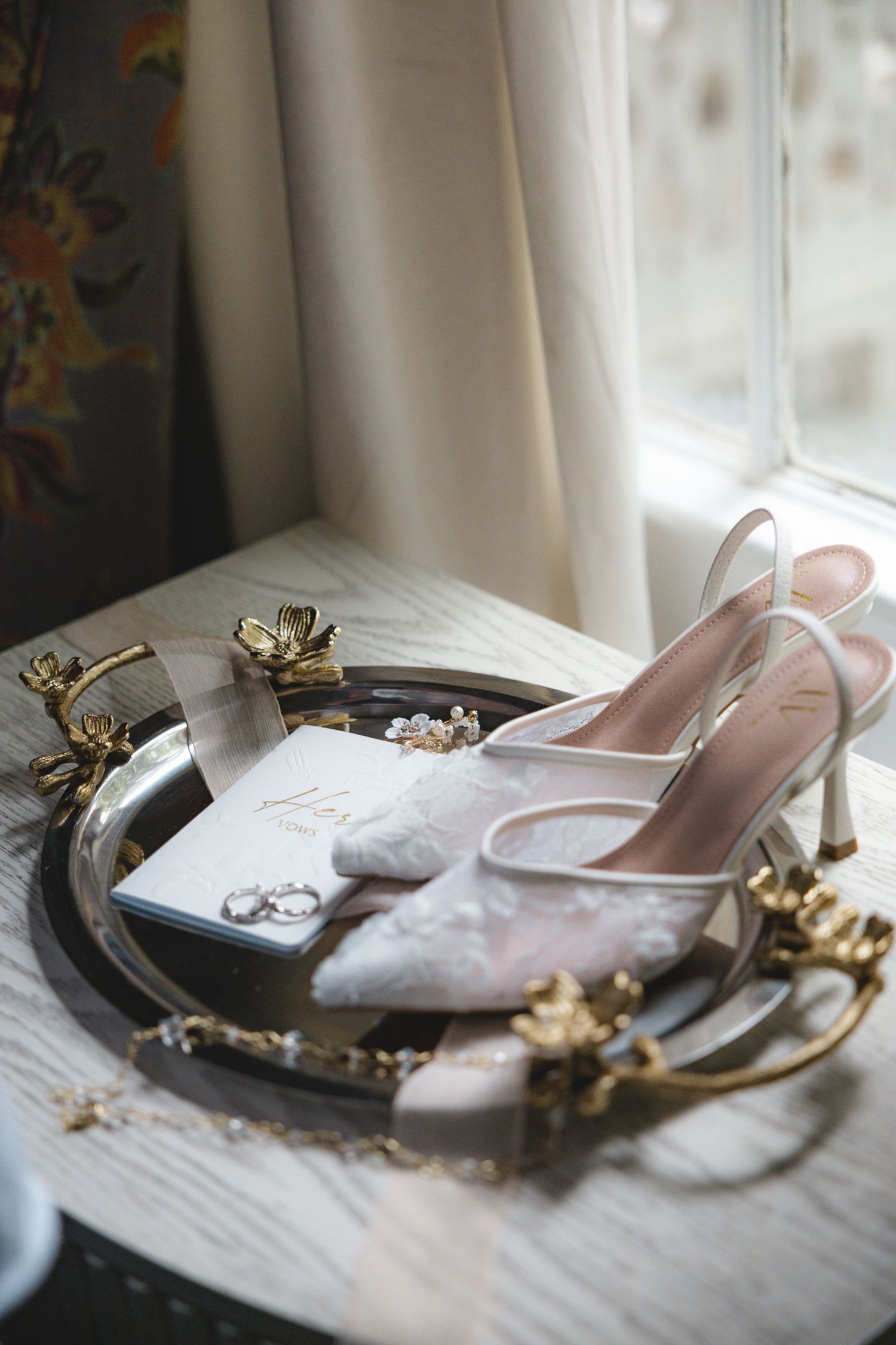 A pair of elegant pink high heeled shoes and jewelry displayed on a wooden surface near a window.