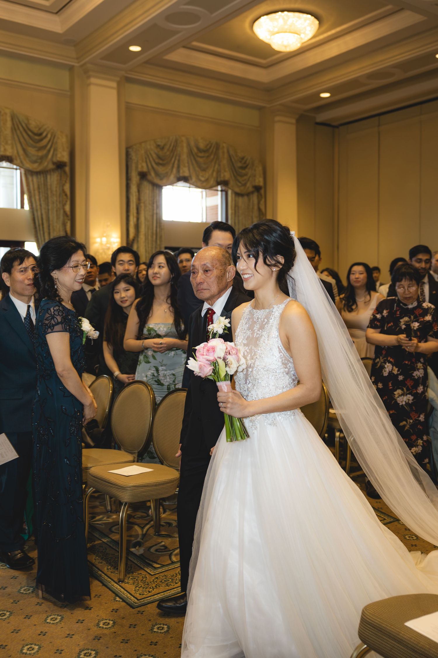 Bride in white gown walks down aisle with father holding pink bouquet in ballroom.