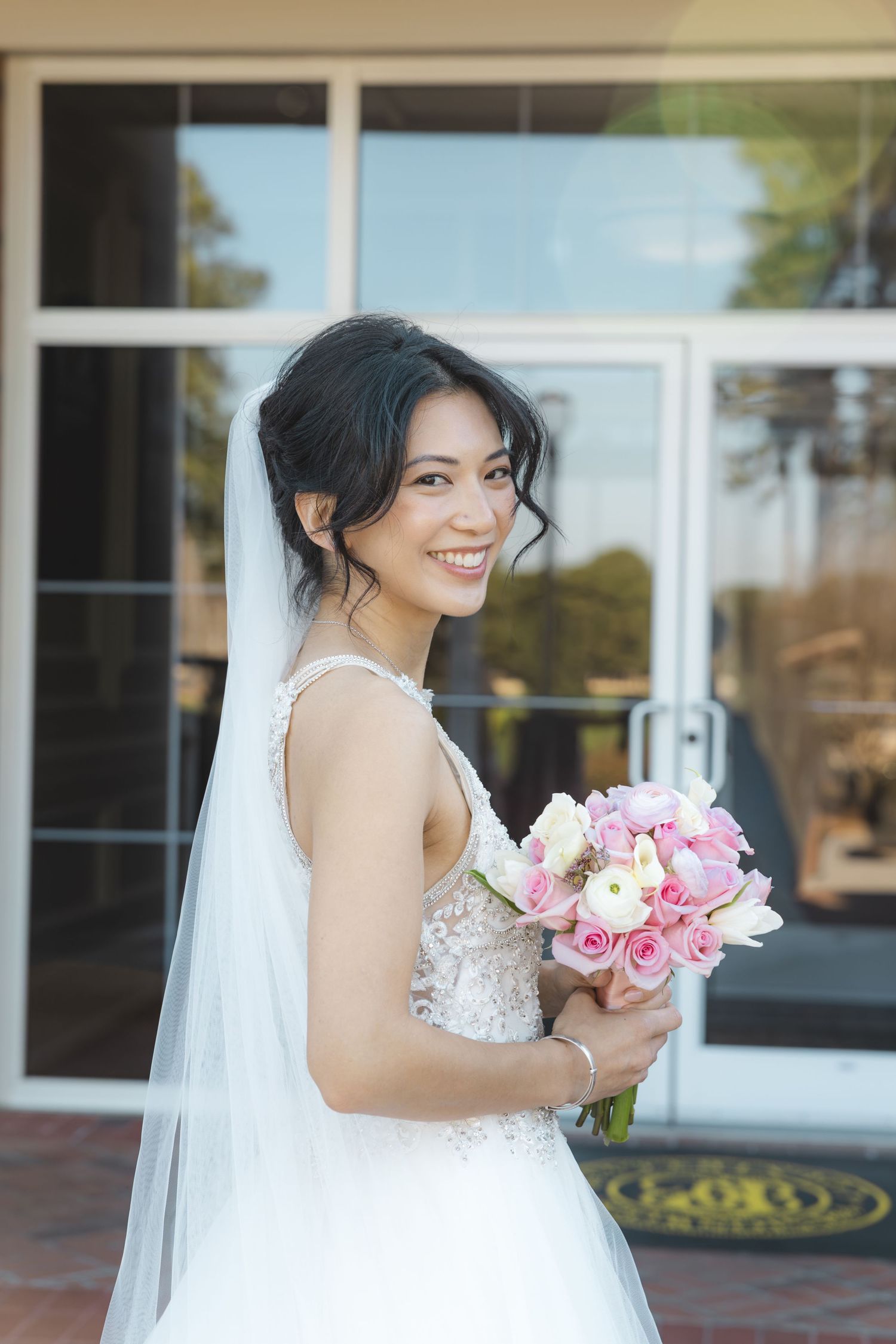 Smiling Asian bride in lace wedding gown holding pink and white bouquet outside venue doors.