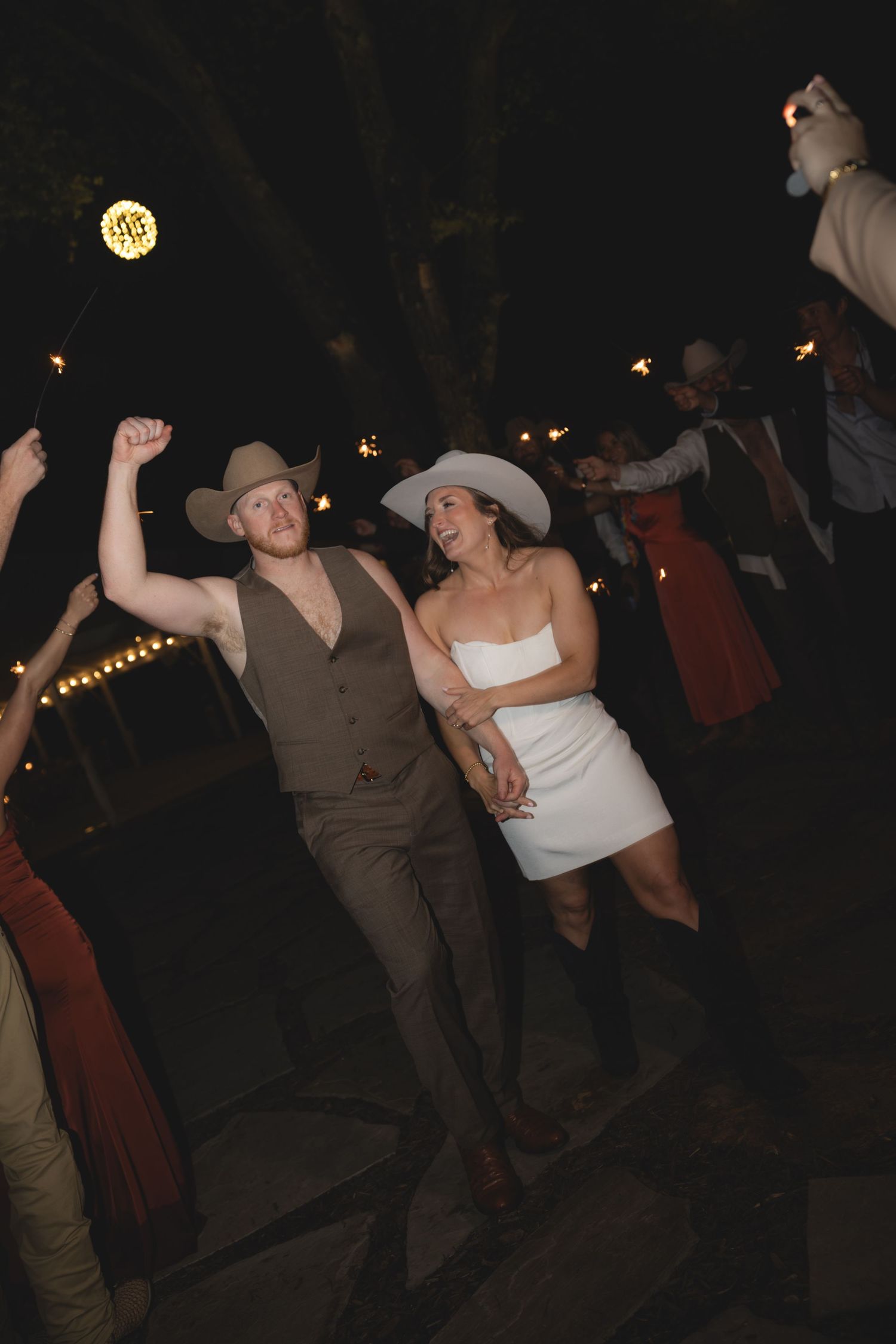 Bride in white mini dress and groom in tan cowboy hat dancing joyfully outdoors at their wedding reception.