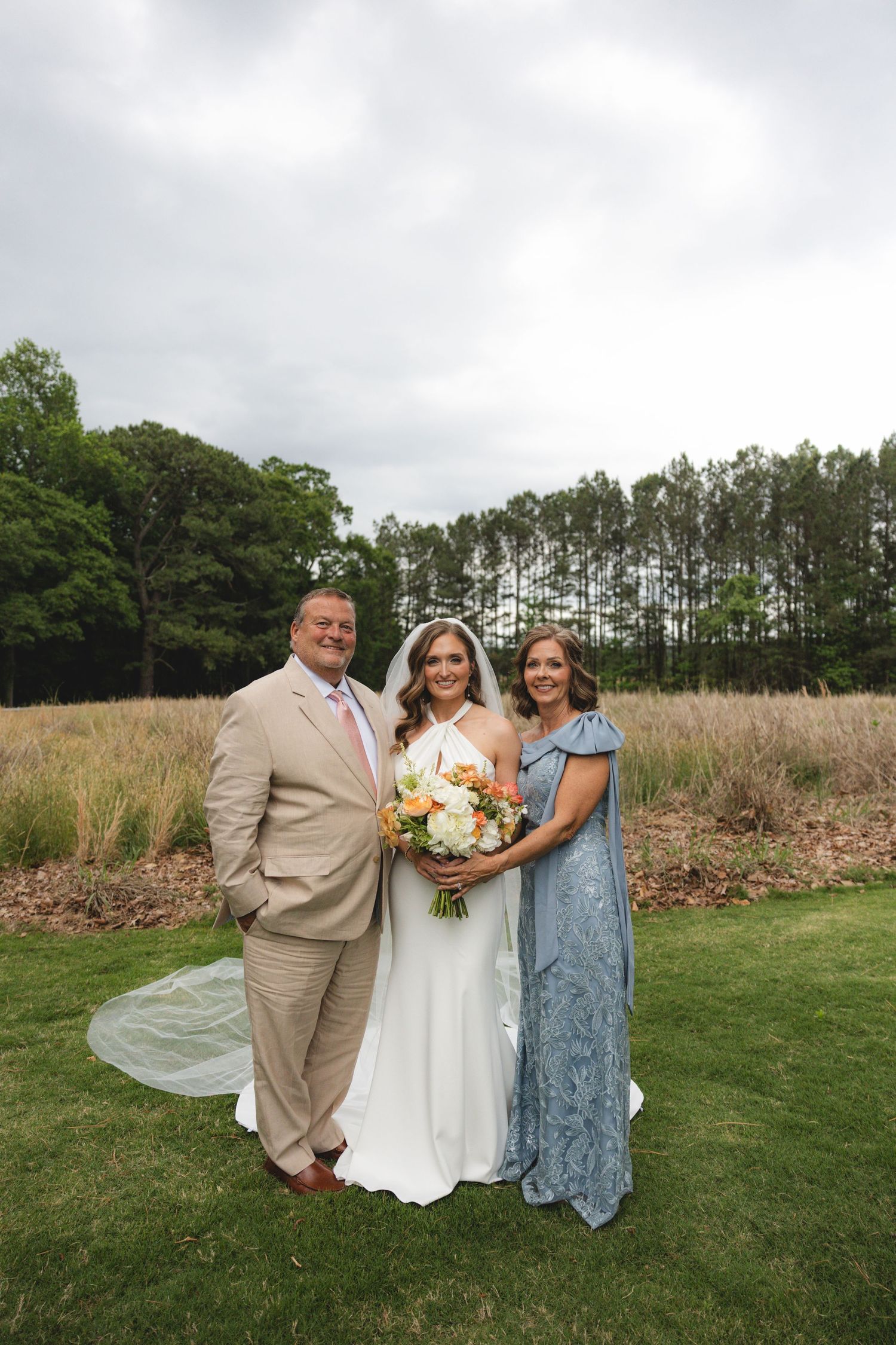 Bride in white halter gown holds bouquet while posing with parents on green lawn at outdoor wedding.