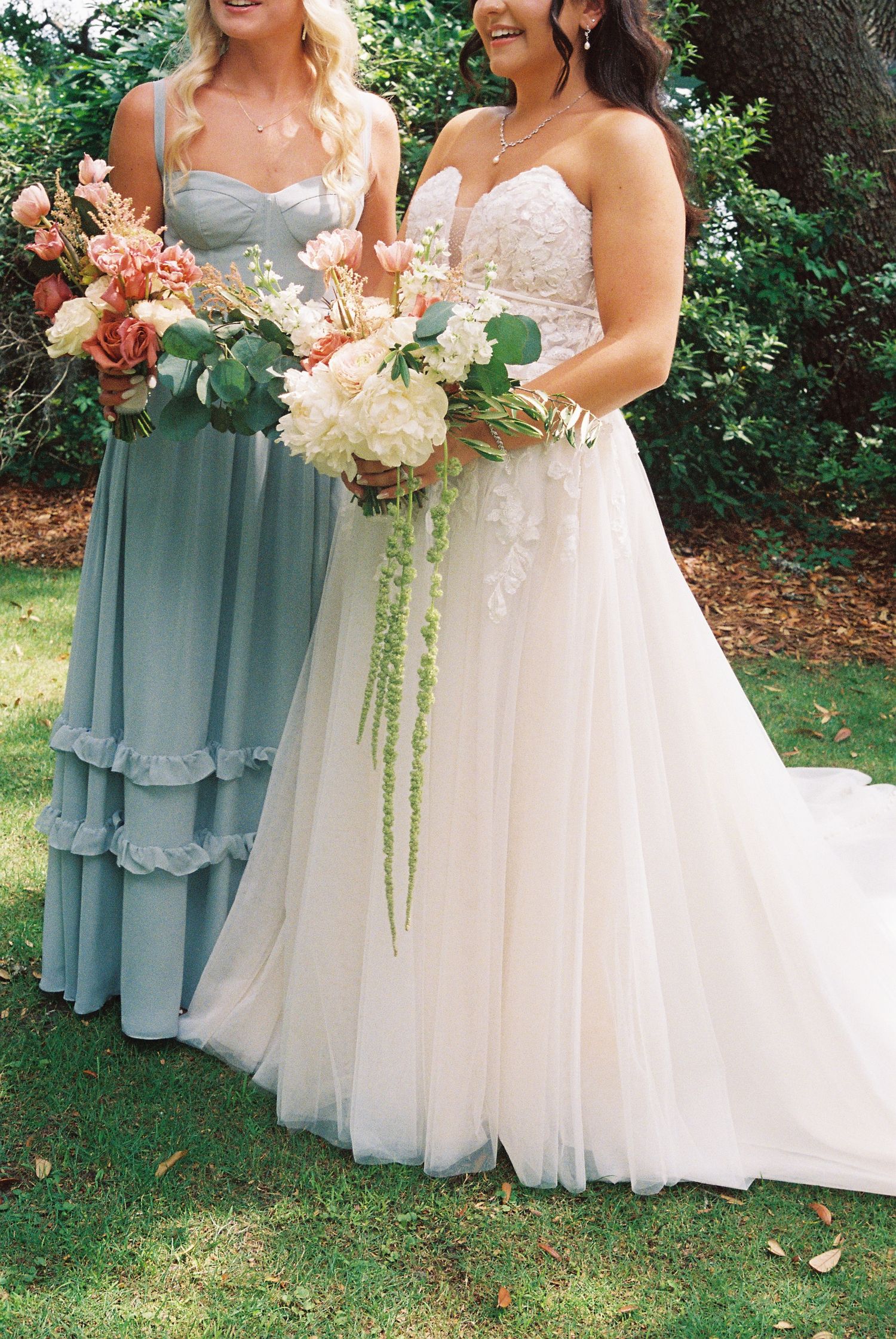 Bride in white lace A-line gown holds floral bouquet beside bridesmaid in dusty blue dress outdoors.