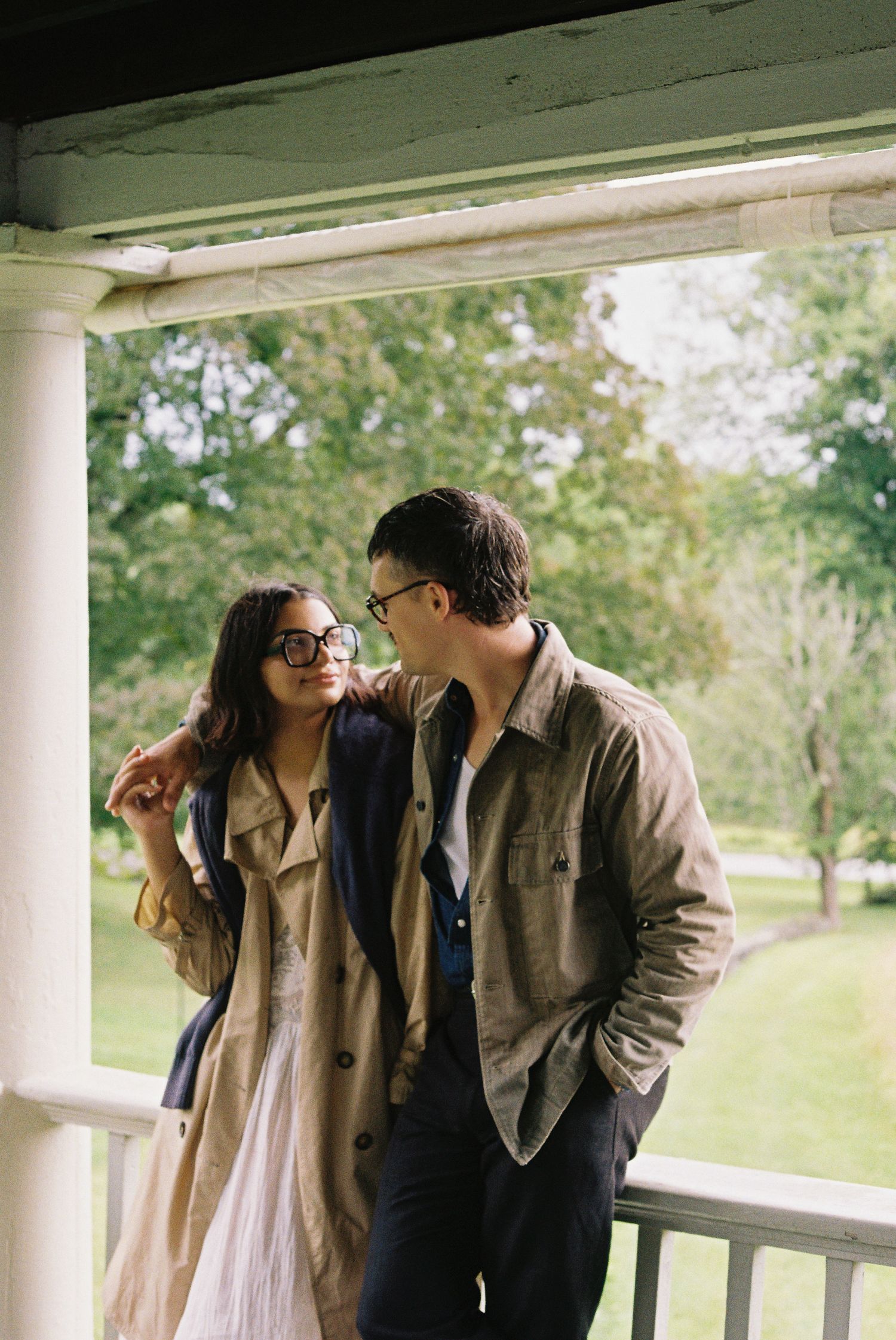A couple wearing glasses shares a tender moment on a porch, surrounded by lush green trees in the background.