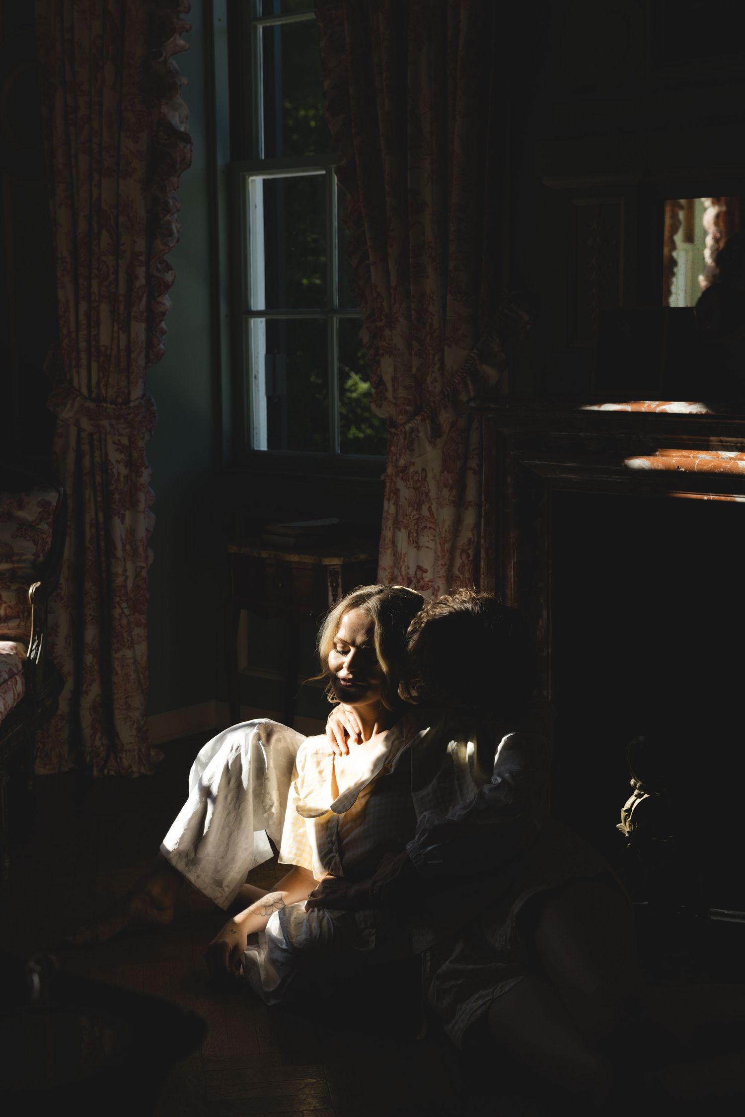Woman in white dress sits in a darkened vintage room, illuminated by soft natural light from an antique window.