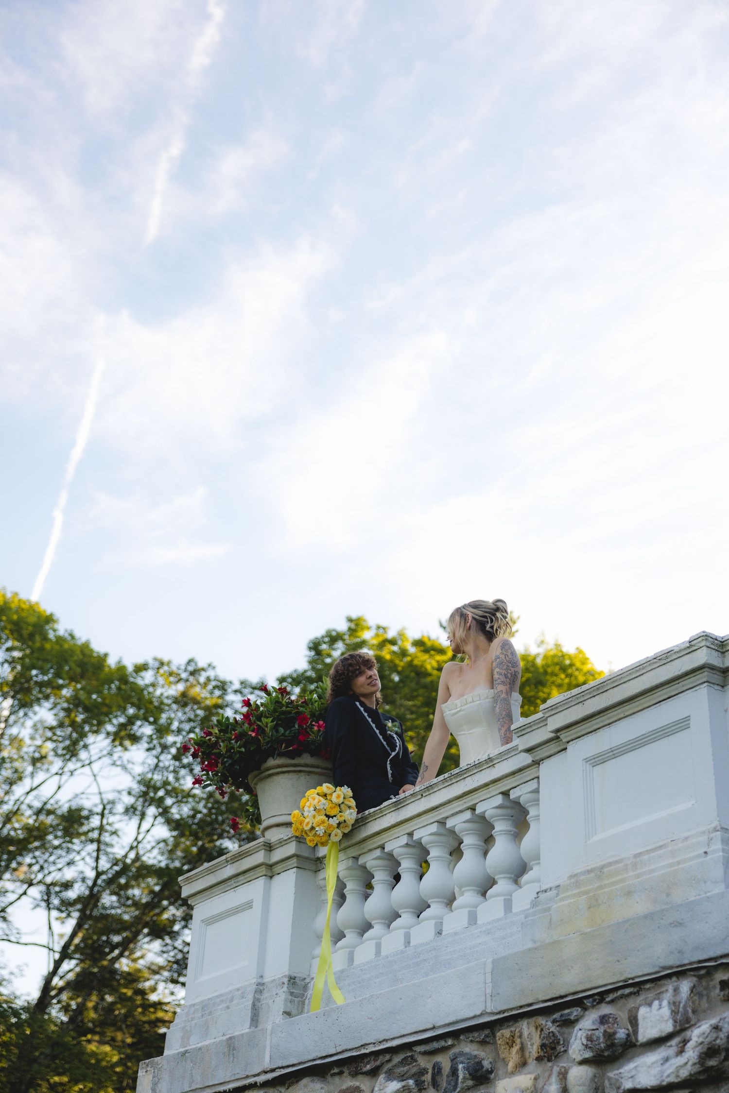 Bride and groom standing on elegant white balustrade balcony with floral arrangements and yellow bouquet against blue sky.