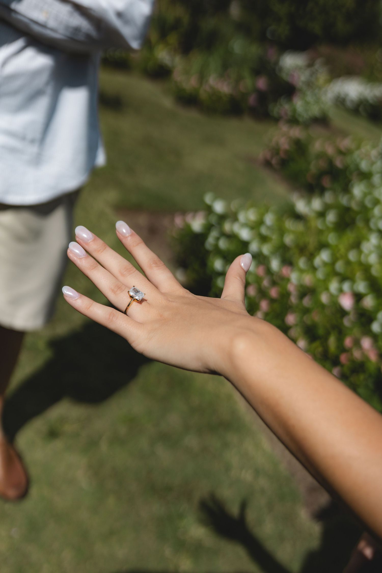 Close-up of a woman's hand displaying a sparkling diamond engagement ring in a lush garden setting.